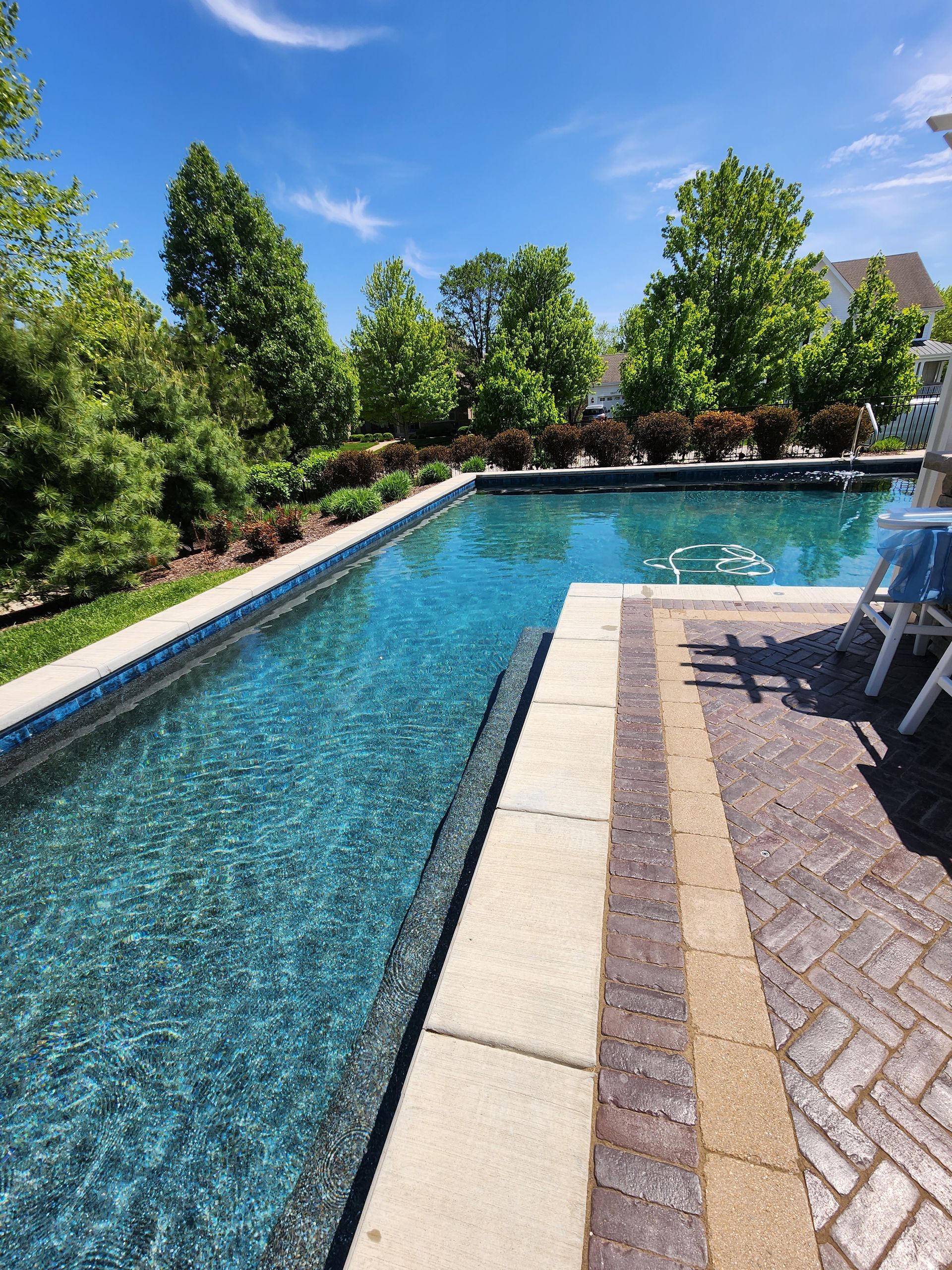A rectangular swimming pool with deep blue water, surrounded by a brick patio, lush green trees, and a clear blue sky.