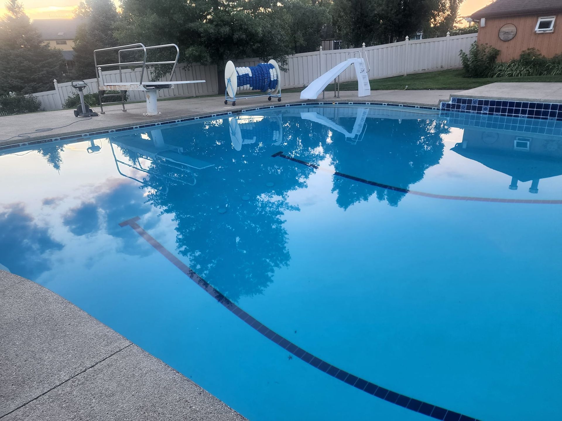 A residential backyard swimming pool at dusk, featuring a diving board, a pool cover reel, and a small slide.