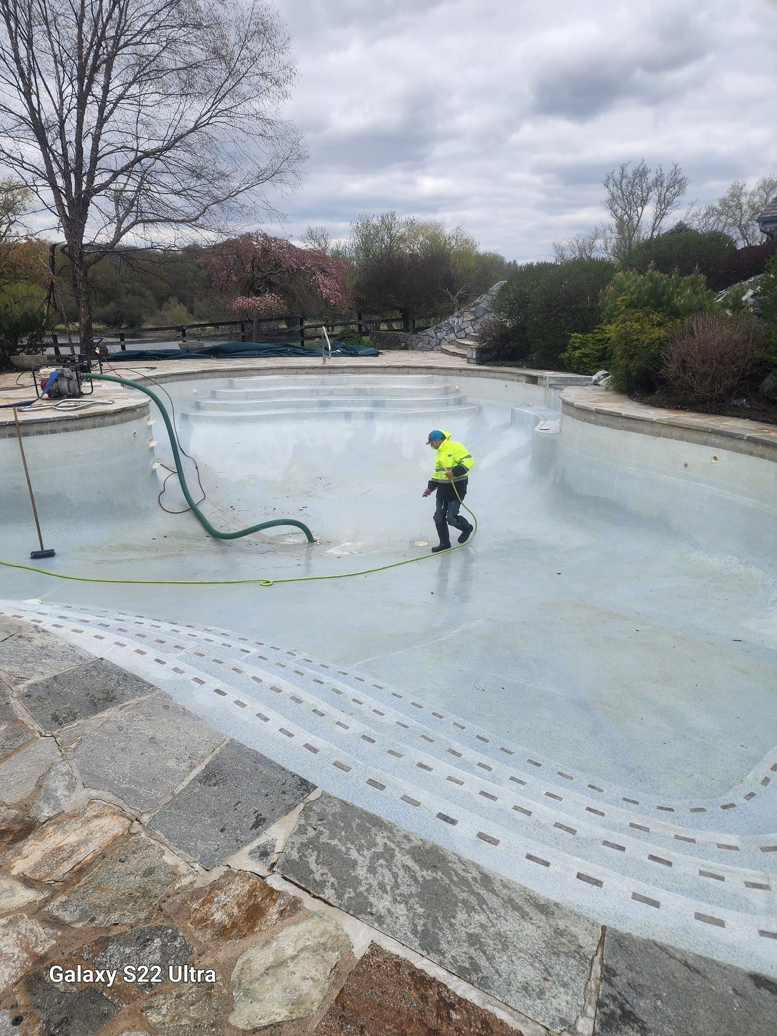 A worker in a high-visibility jacket pressure washes the surface of an empty, light-blue residential swimming pool.