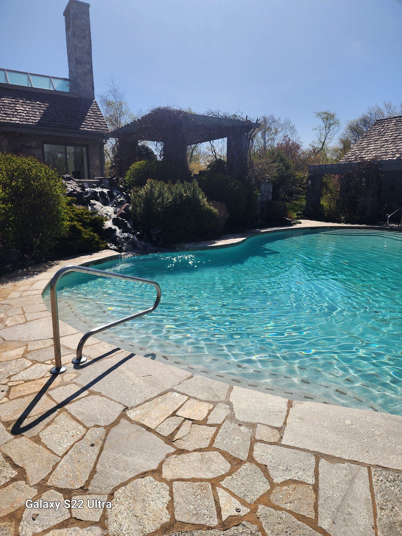 A stone pool deck with a metal handrail overlooks a turquoise swimming pool featuring a waterfall and a pergola.