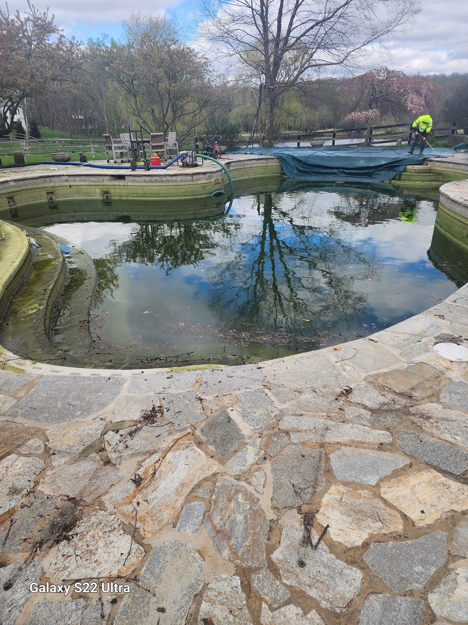 A stone patio overlooks a murky pond reflecting trees and sky, with a small bridge and a cyclist in the distance.