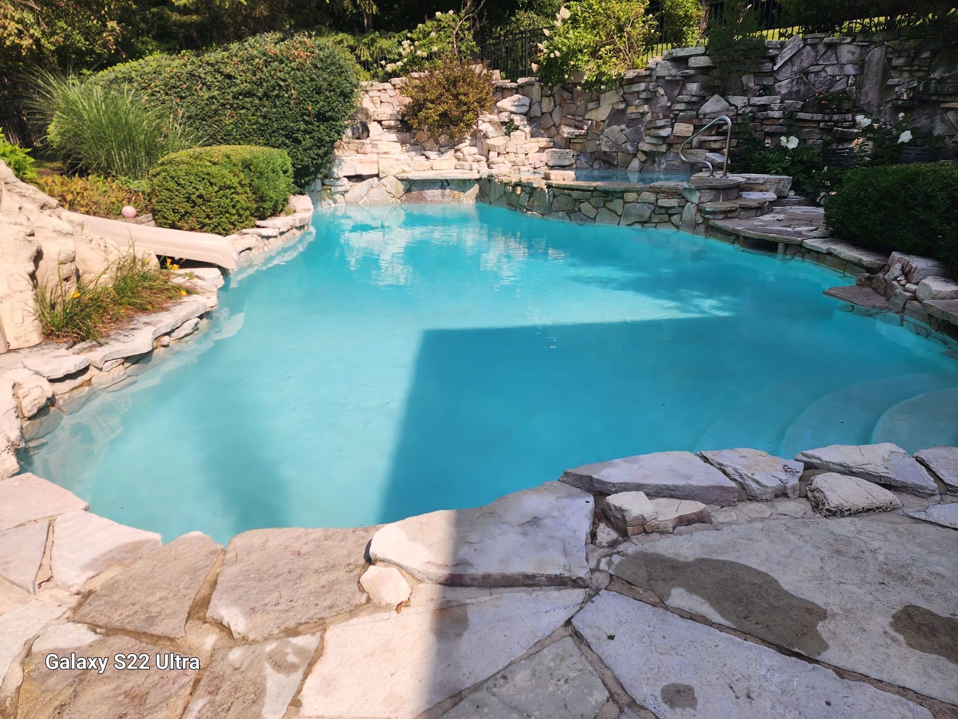 A turquoise swimming pool framed by natural stone decking and surrounded by lush green bushes and stone landscaping.