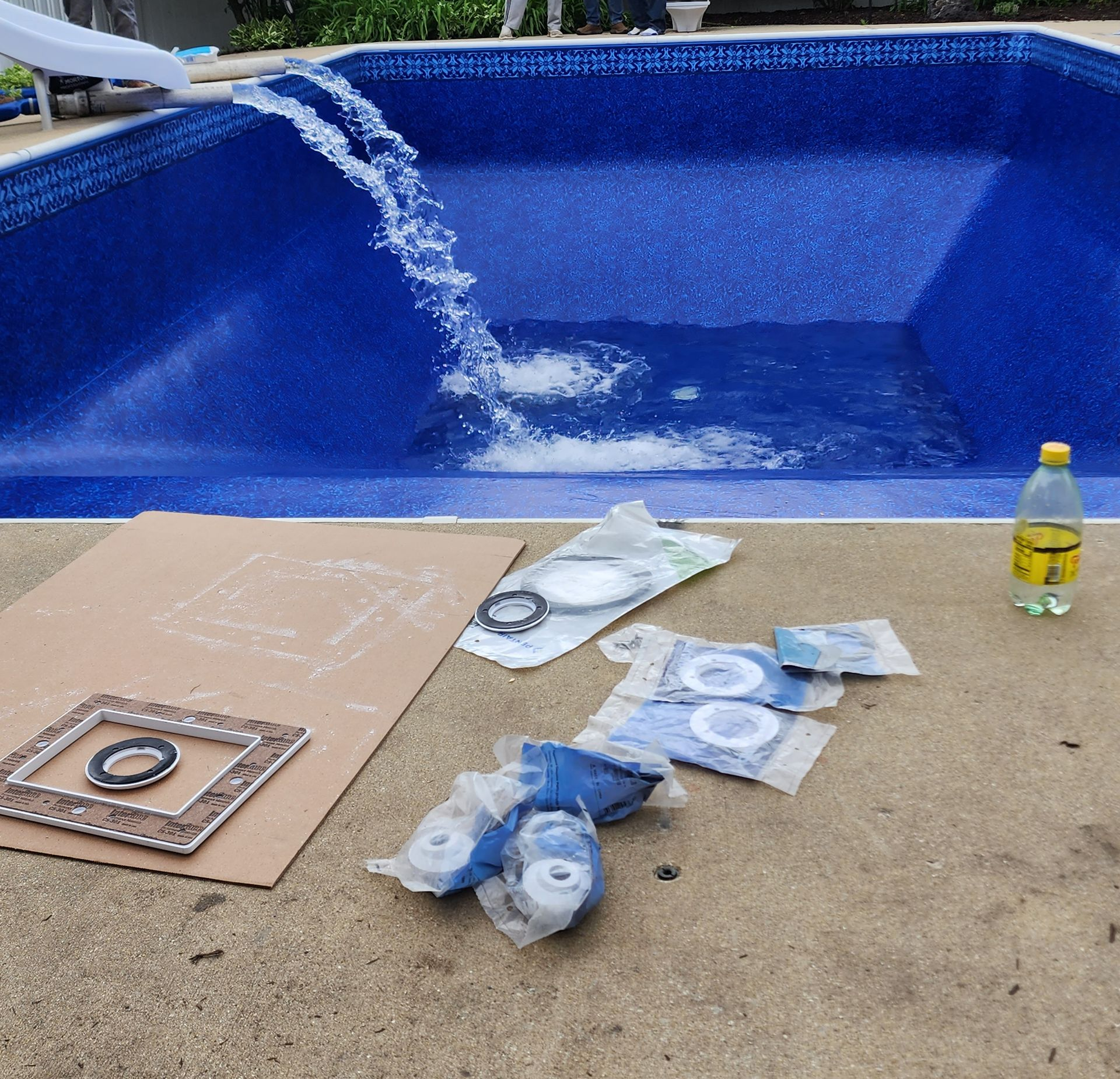 A hose fills a blue, empty swimming pool while pool replacement parts lie on the concrete patio nearby.