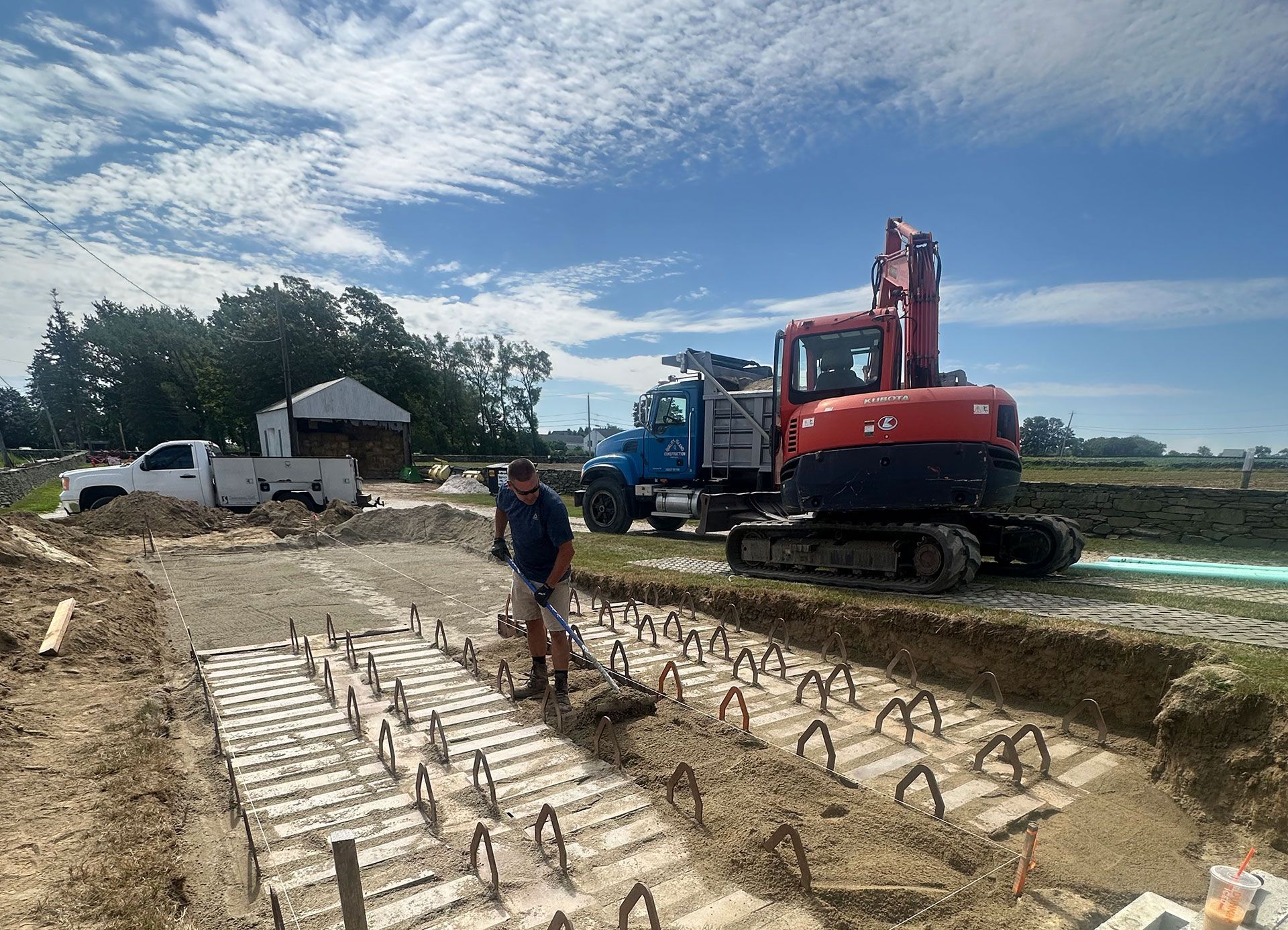 A person works on a construction site with rows of steel rebar protruding from concrete footings, an excavator, and trucks.