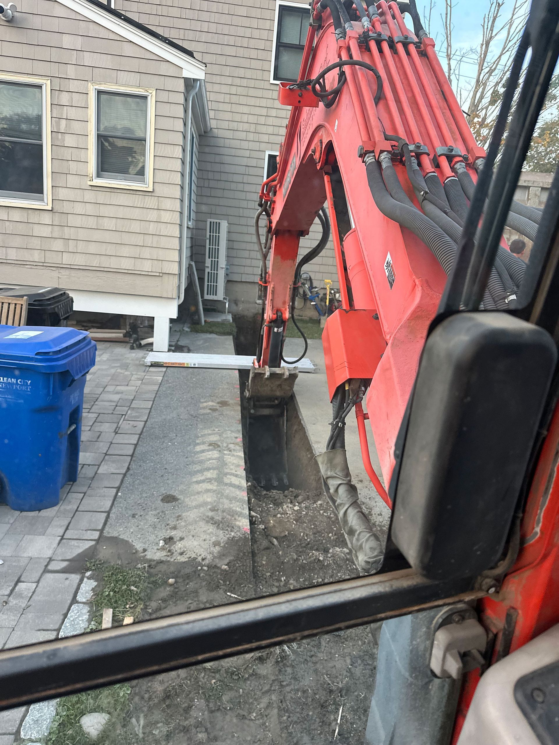 A view from the cab of an orange excavator digging a narrow trench alongside a house next to a blue trash bin.