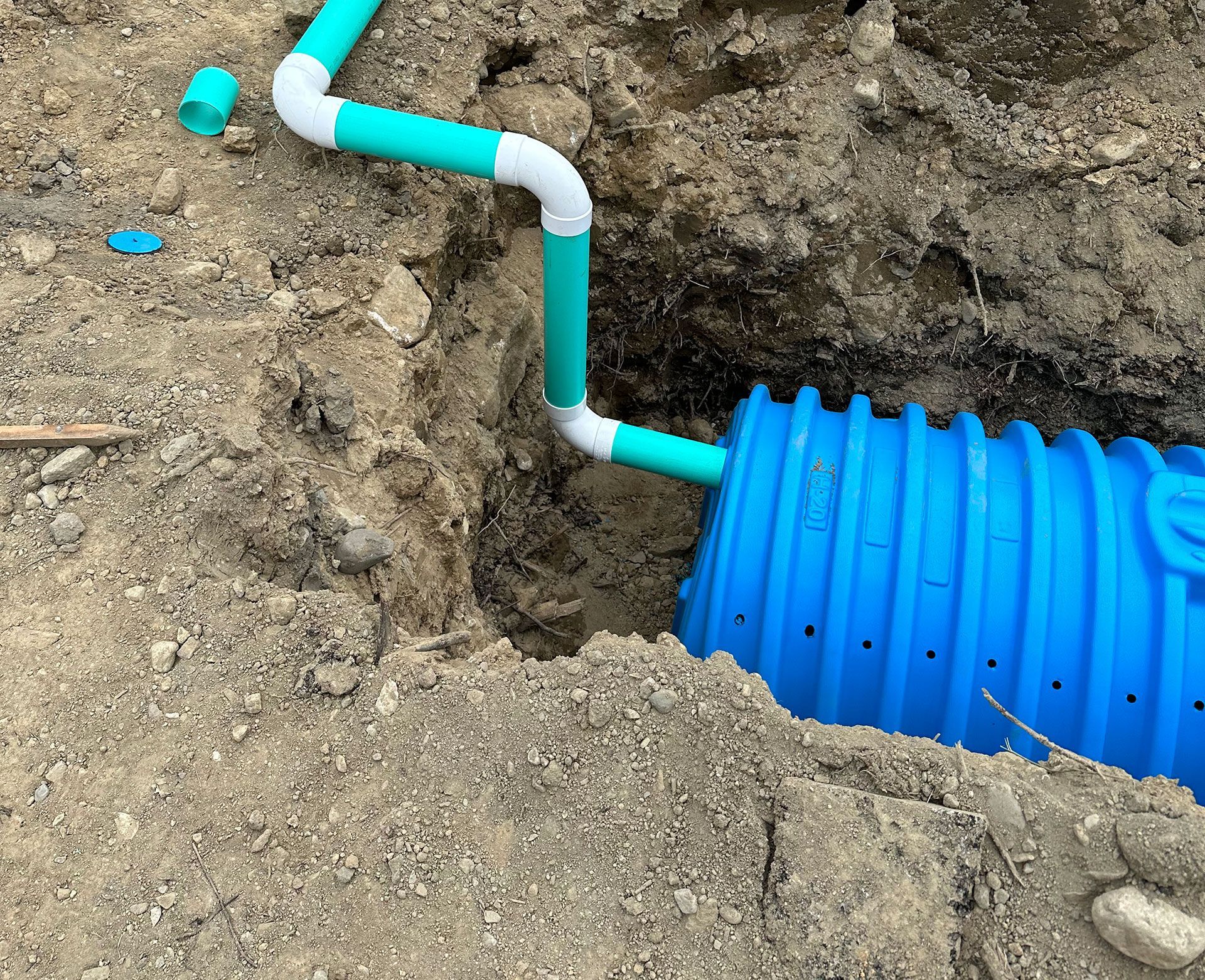 A blue plastic septic or drainage chamber partially buried in the ground, connected to white and green PVC pipes.
