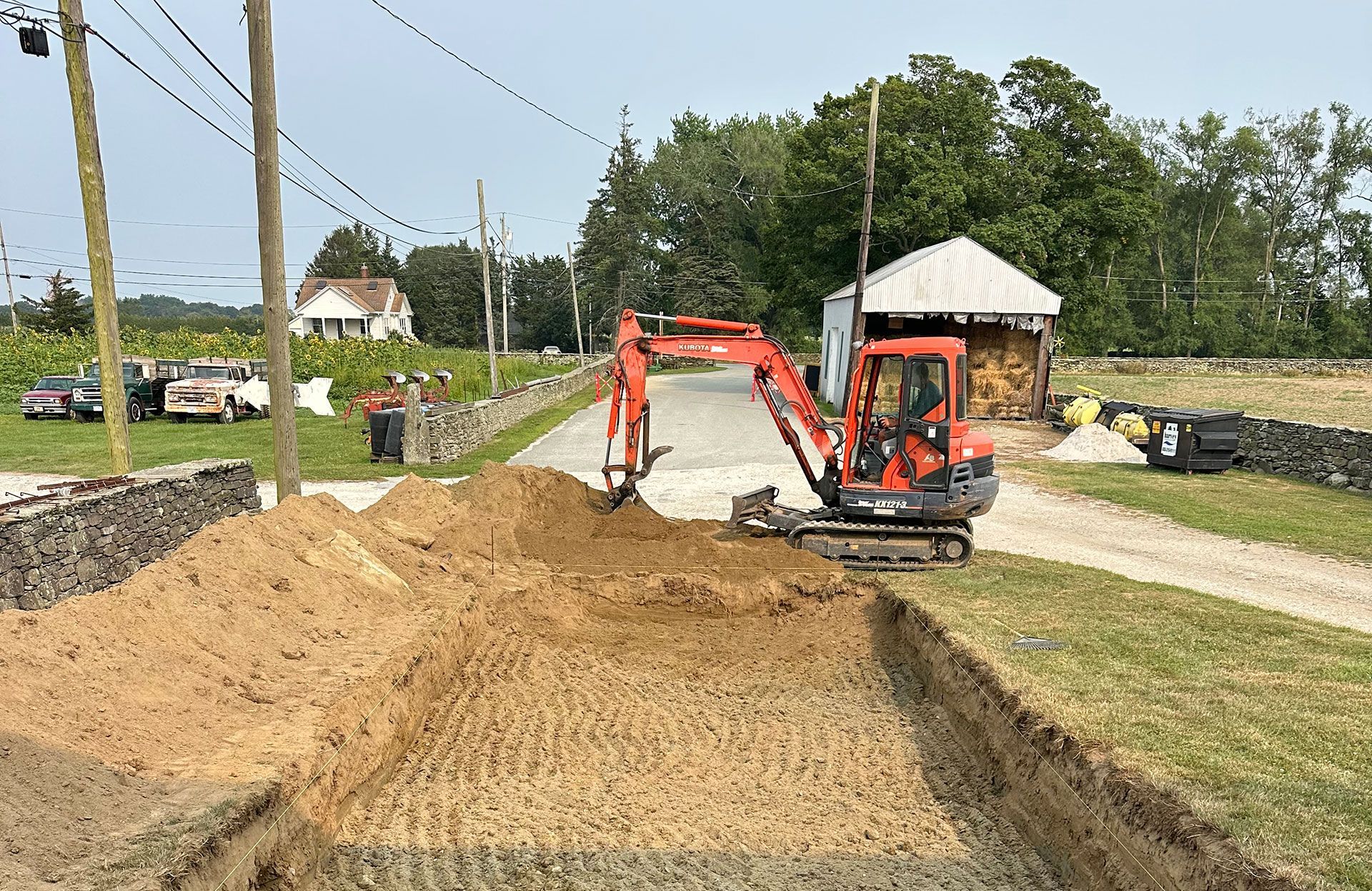 An orange mini-excavator digs a trench in a rural, grassy area near a dirt road, gravel driveway, and farm structures.