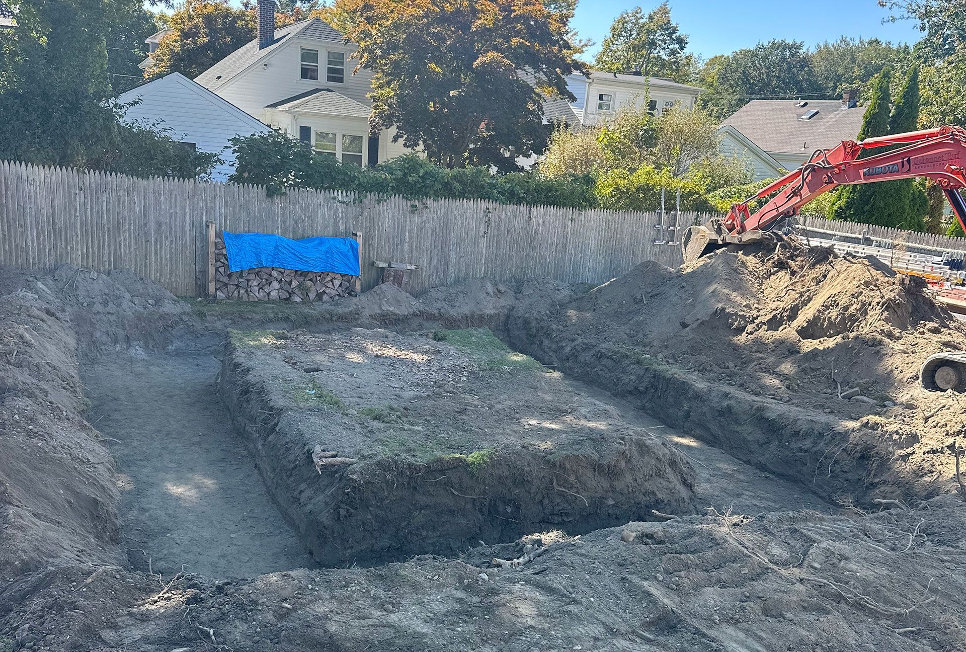 An excavation site with a dirt foundation pit, a wooden fence, and a red excavator arm visible in the background.