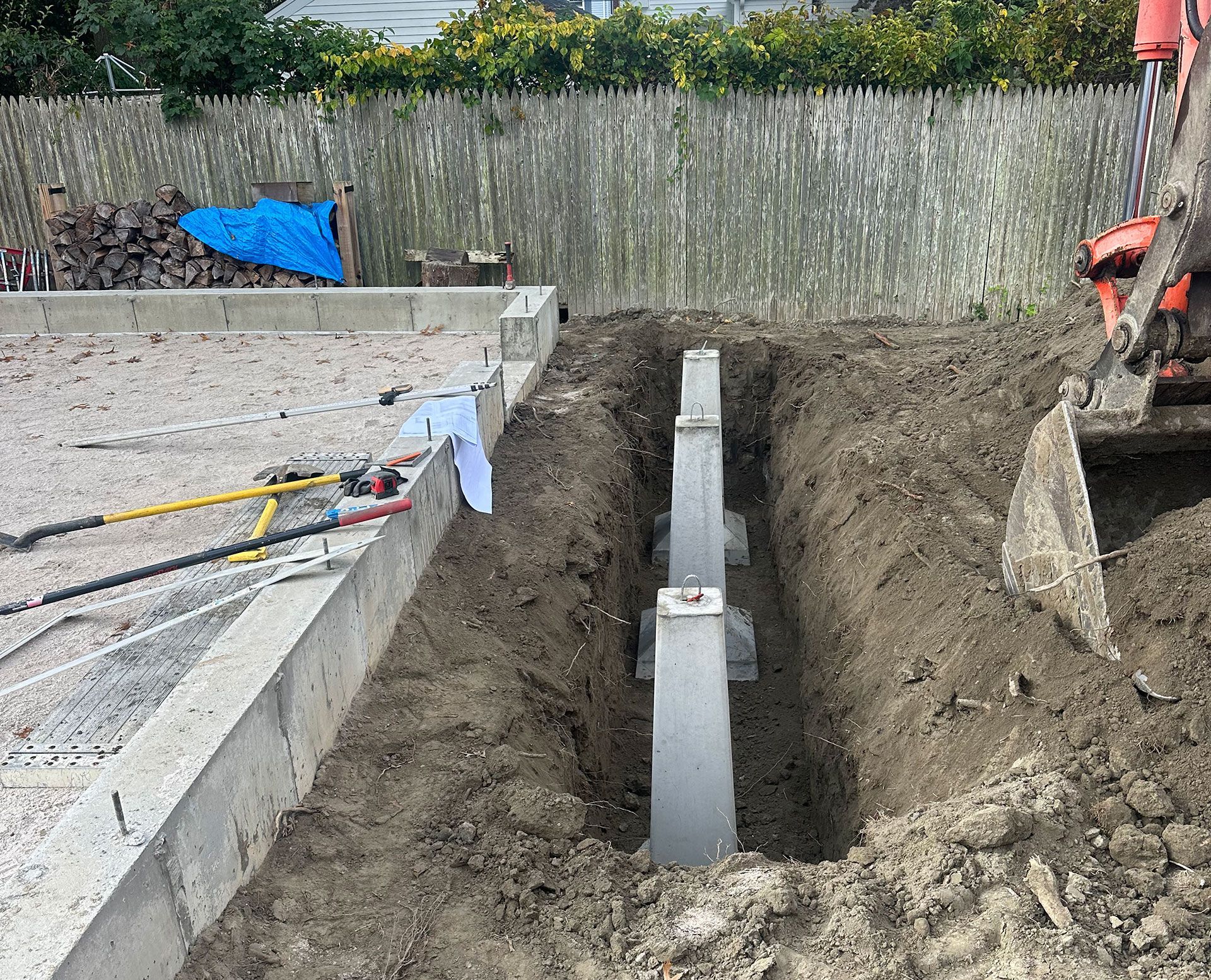 A trench with concrete pier foundations alongside a gravel area and a wooden fence, with an excavator bucket visible.
