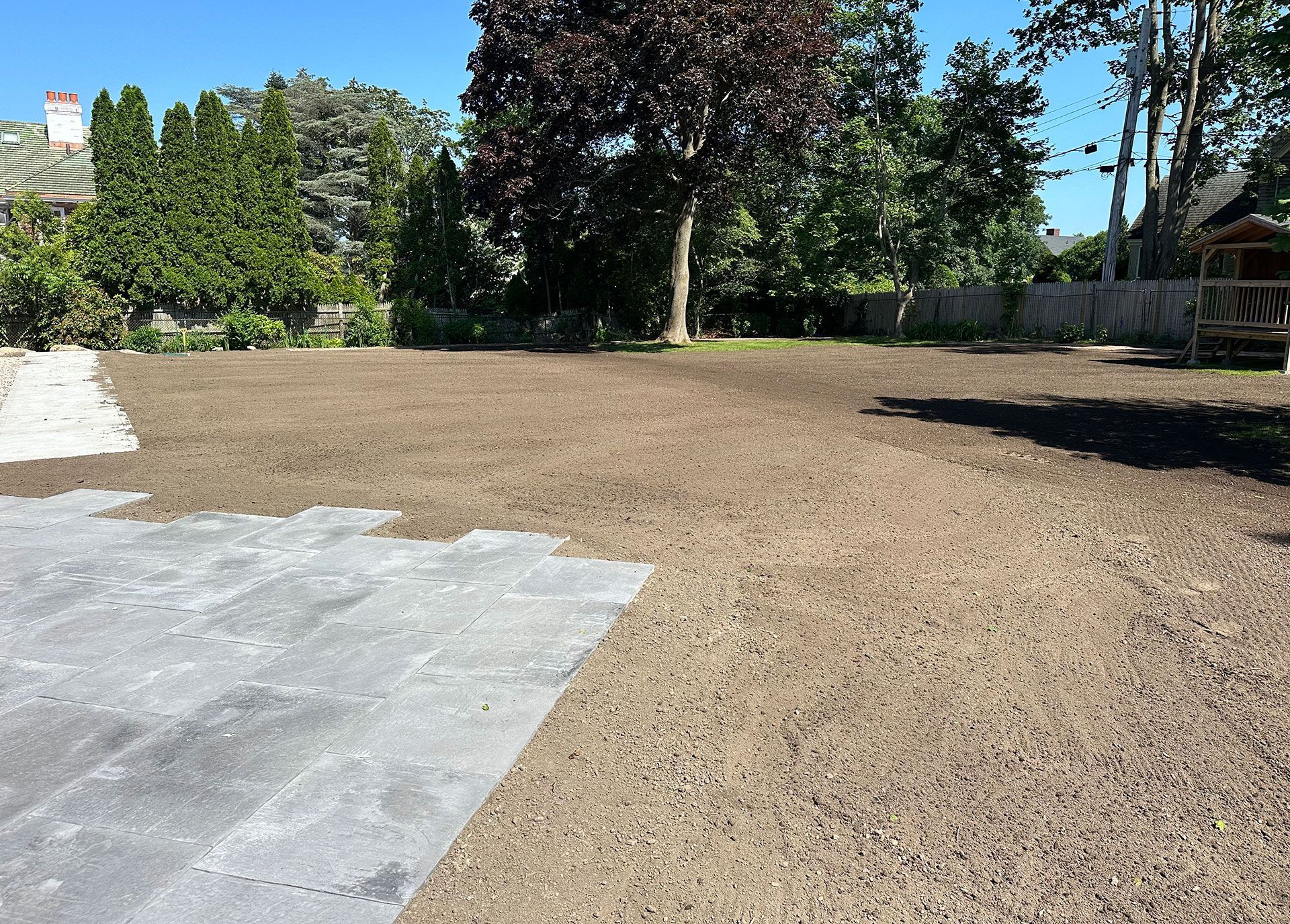 A backyard area featuring a large, freshly prepared dirt lawn adjacent to a light gray stone patio under a clear blue sky.
