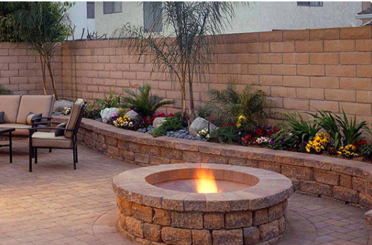 Backyard patio with a stone fire pit, seating, and a landscaped retaining wall at dusk
