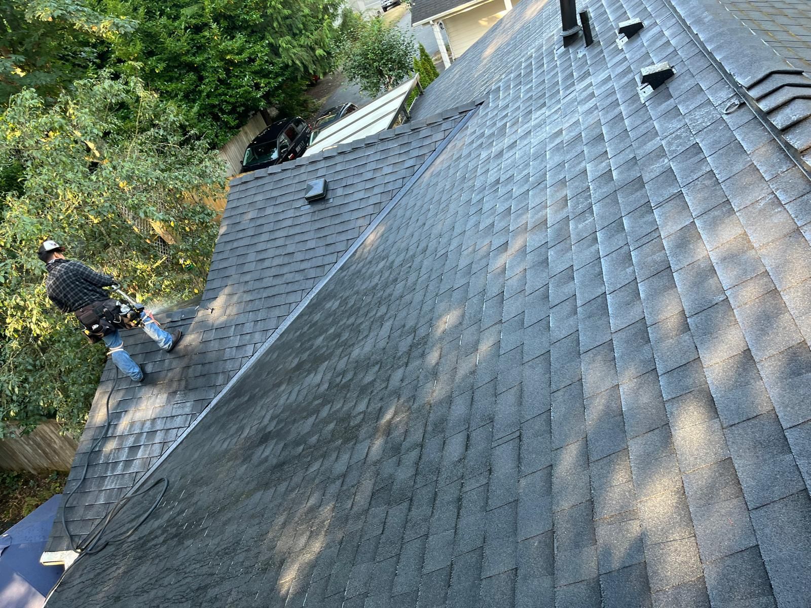 Roofer on a dark shingled roof, trees and other buildings in the background.
