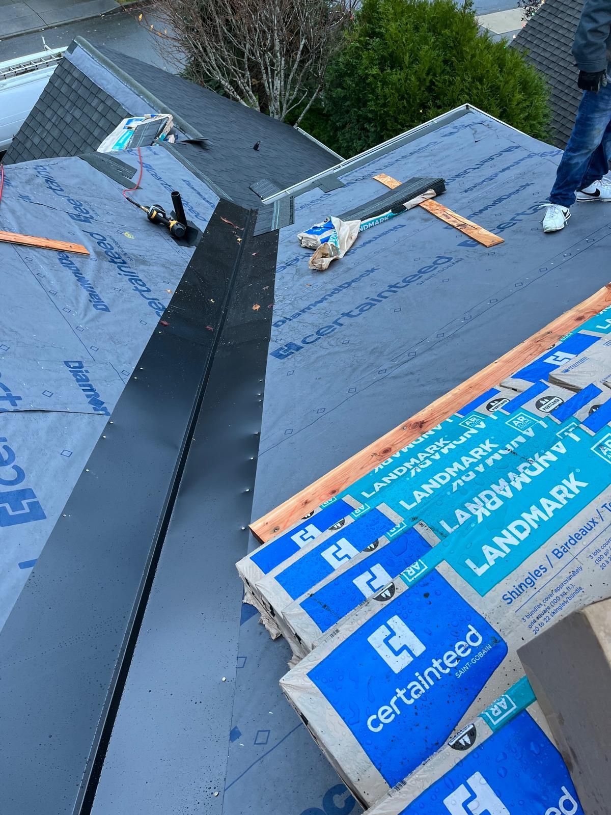 Roofing materials on a roof with a metal flashing. Blue tarp, wooden boards, and bags of shingles are present.