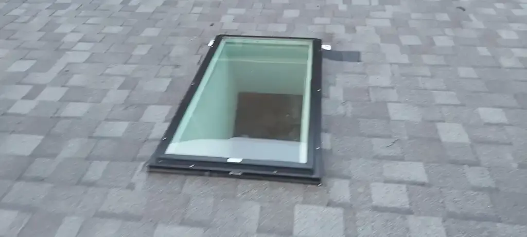 Skylight on a gray shingle roof. The open skylight has a rectangular frame and a glass panel.