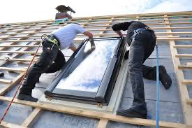 Two workers installing a skylight on a rooftop, wearing safety harnesses.