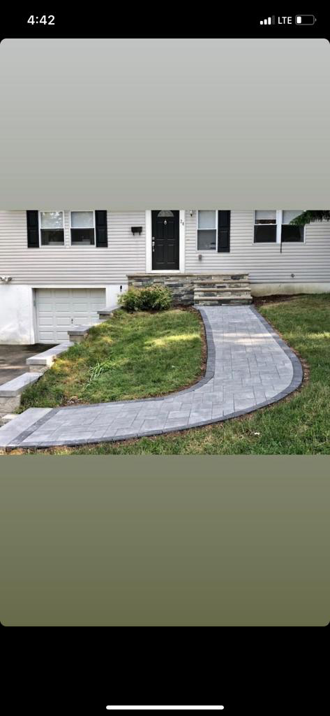 A paved walkway curves up to a house with a black door and shutters. Lush green grass surrounds the path.