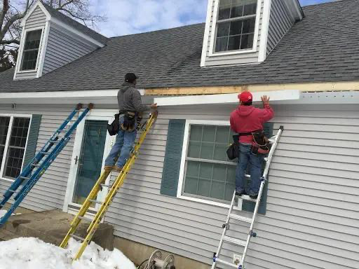 Two workers on ladders installing siding on a house with gray siding and roof.