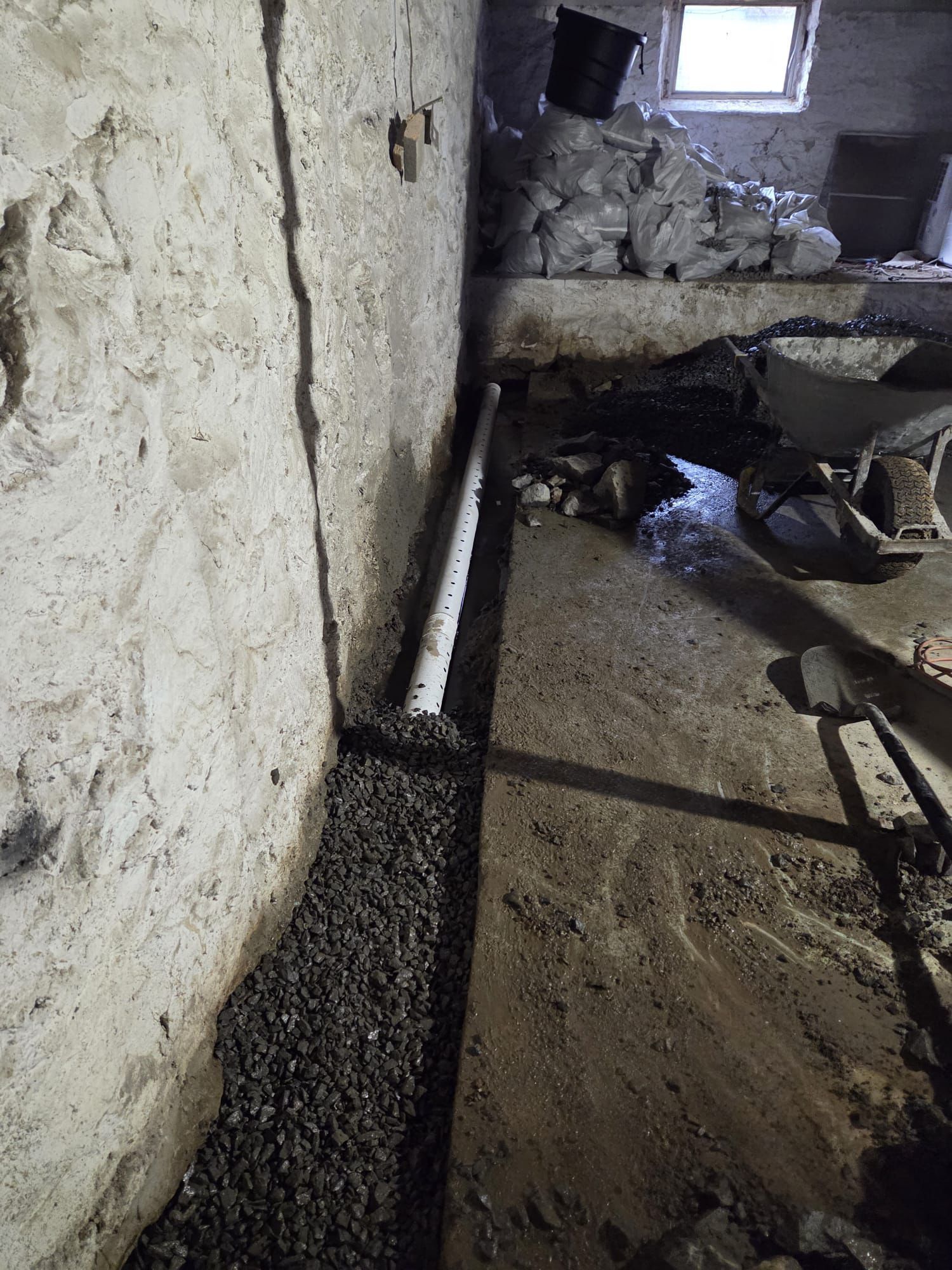 Basement interior with drainage system construction. White pipe in gravel-filled trench along a concrete wall.
