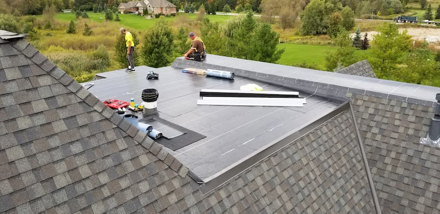 Two roofers on a roof installing dark solar panels, surrounded by asphalt shingles and a green landscape.
