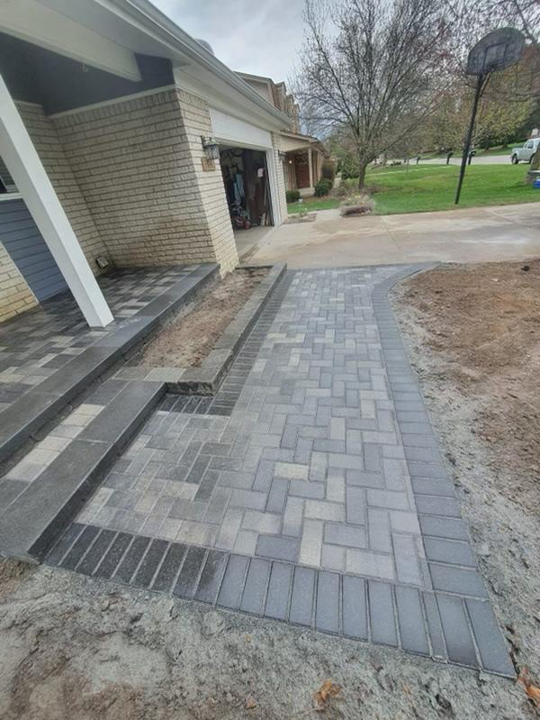 Brick pavers create a walkway and steps leading to a house entrance. Gray and black bricks are used.