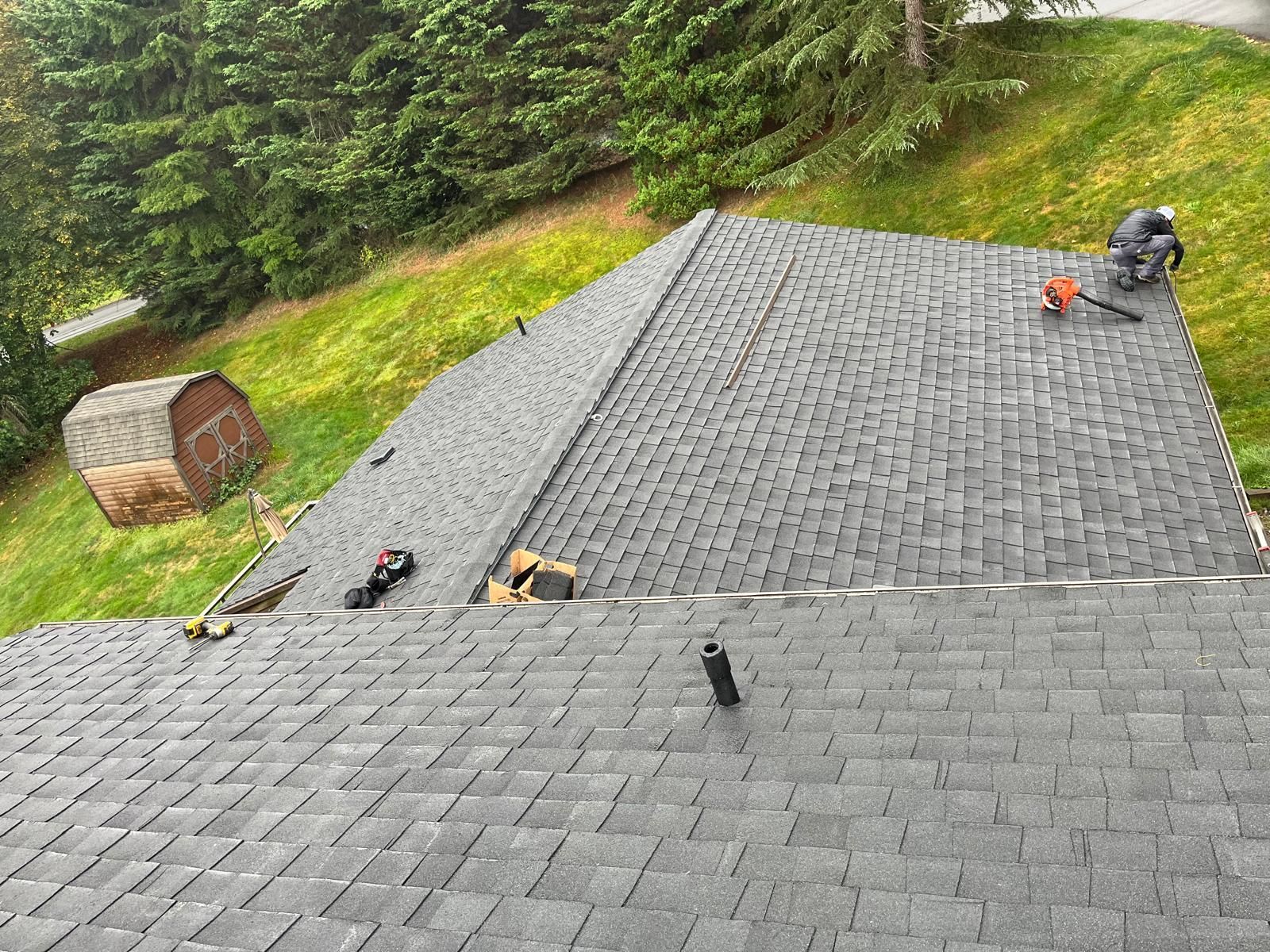 Roofer on a gray shingled roof, tools nearby. Shed and green grass in the background.