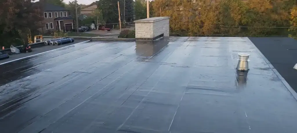 A wet, black rooftop with a small building, chimney and trees in the background.