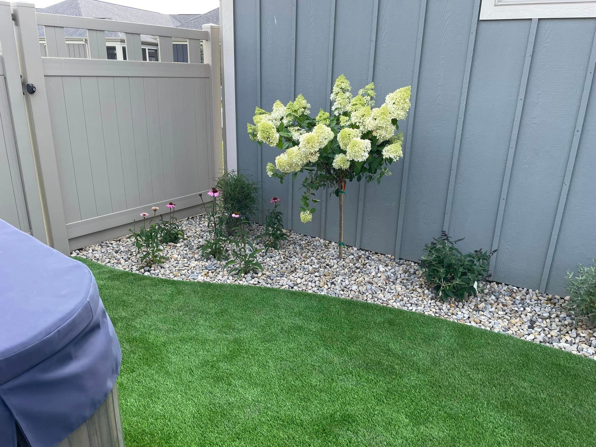A backyard with a fence , grass , flowers and rocks.