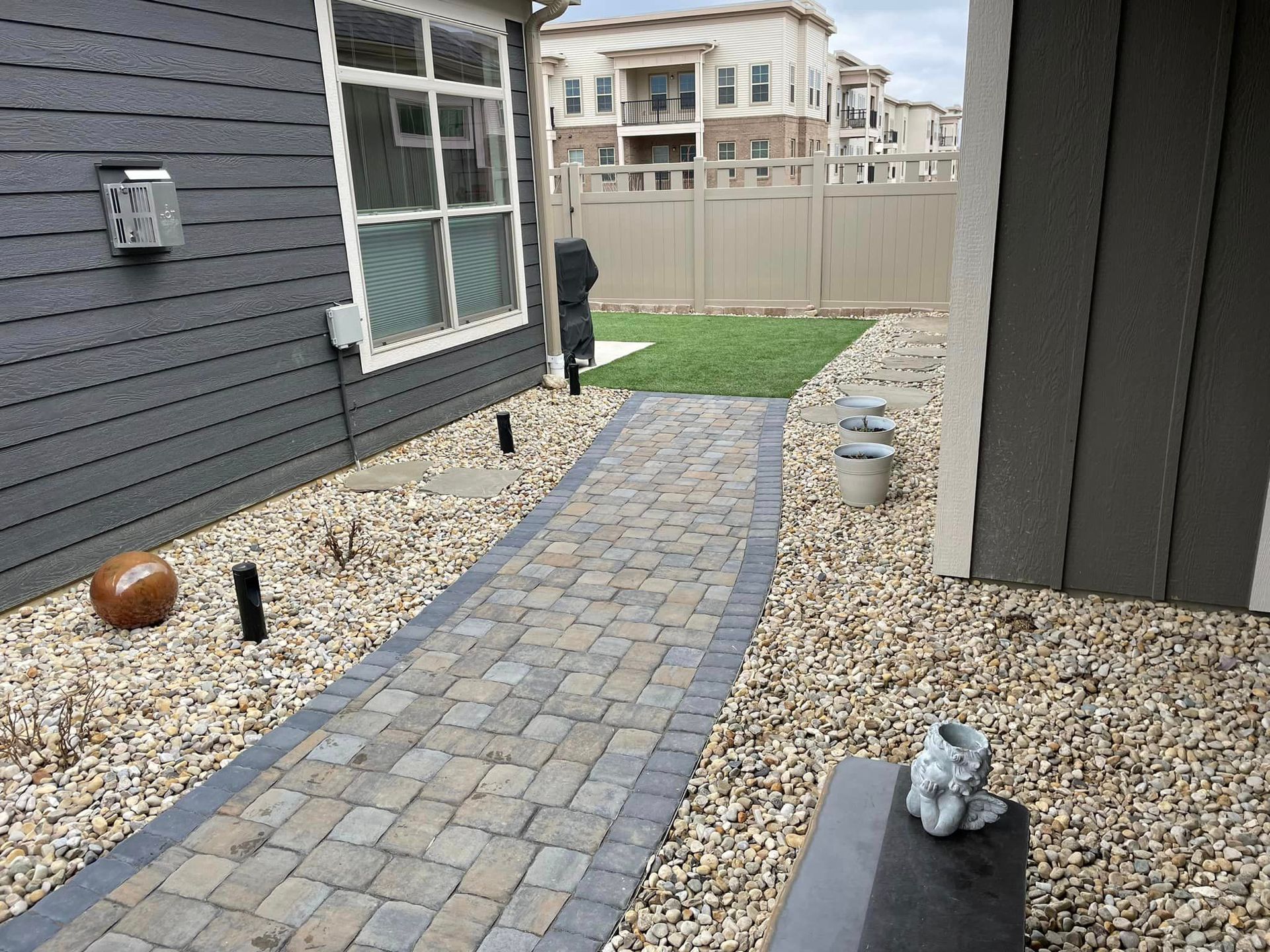 A brick walkway leading to a house with a fence in the background