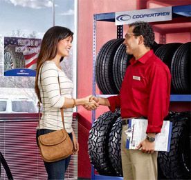 Woman shaking hands with a tire shop employee beside stacked tires and a Cooper Tire display