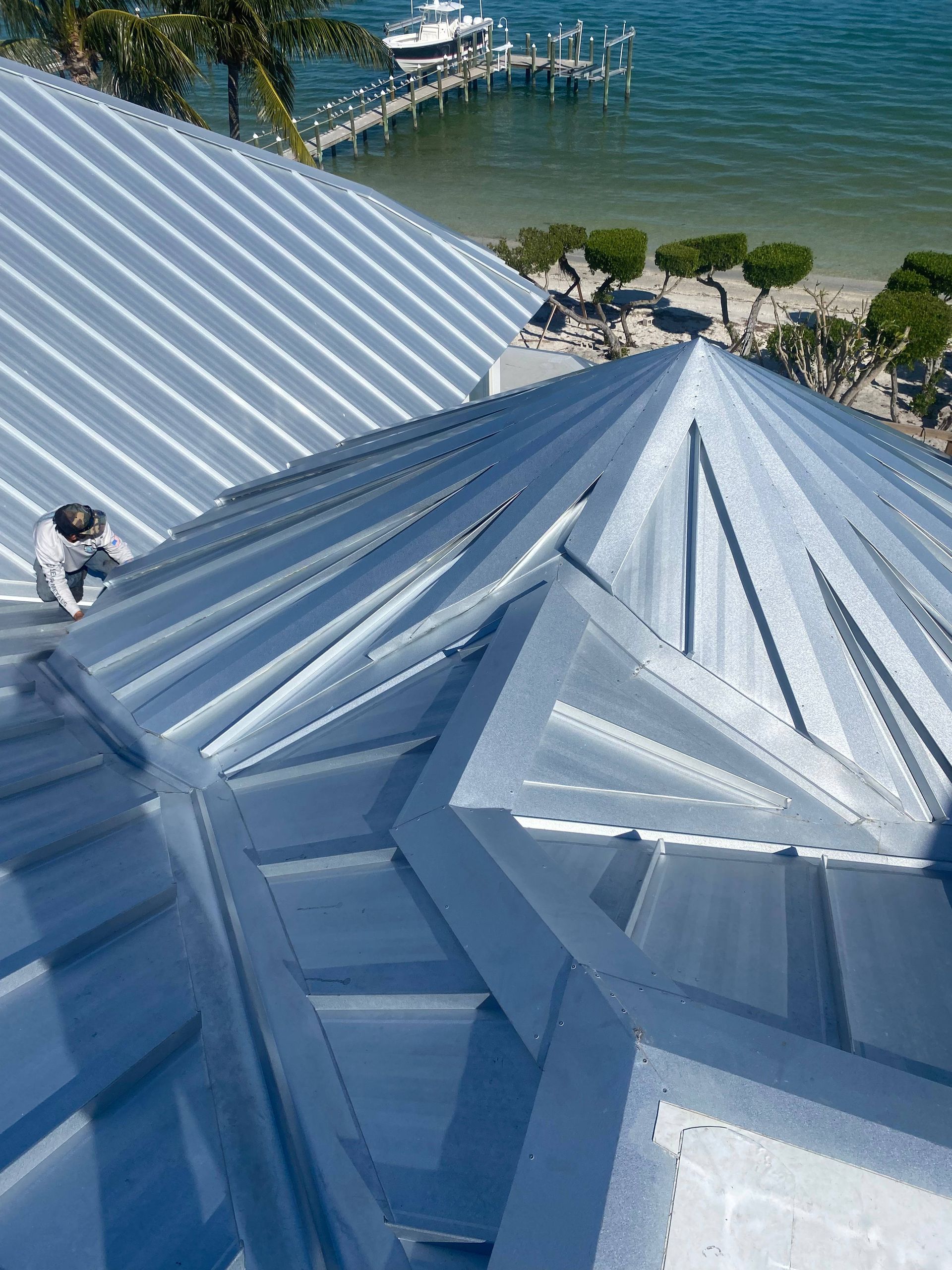 Worker on a silver metal roof with a view of a pier and water in the background.