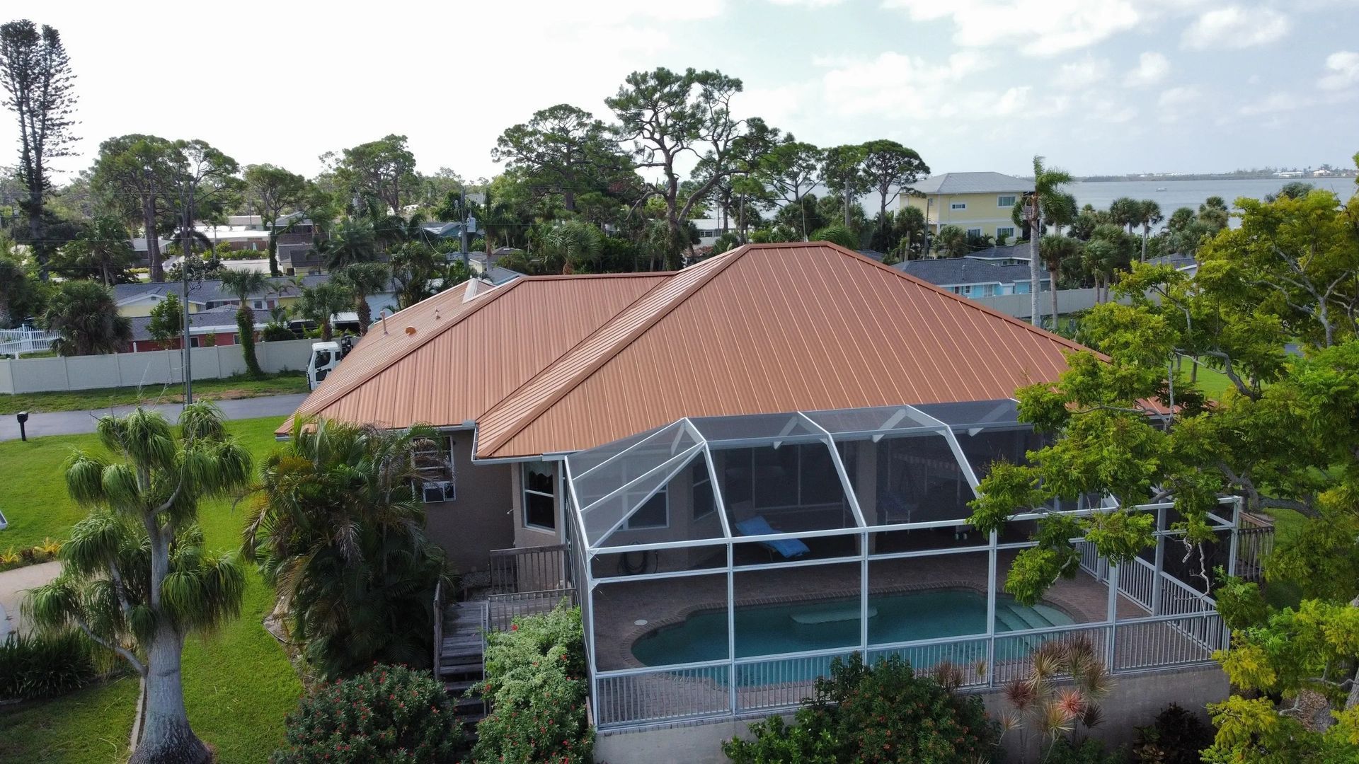 House with brown roof, screened pool enclosure, surrounded by green trees and grass, near water.