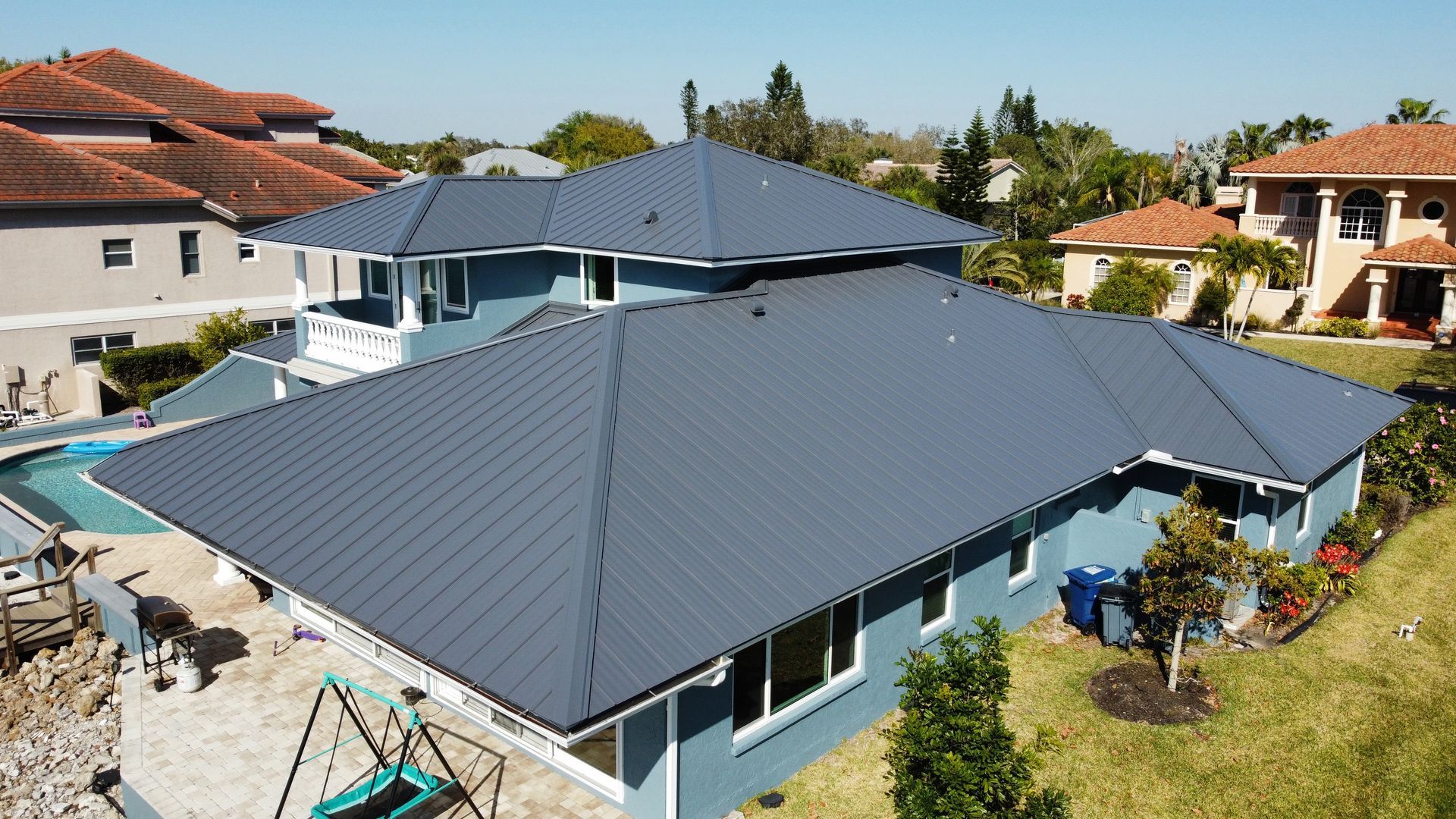 Blue roof on a large house, surrounded by other houses with orange roofs, near a pool.