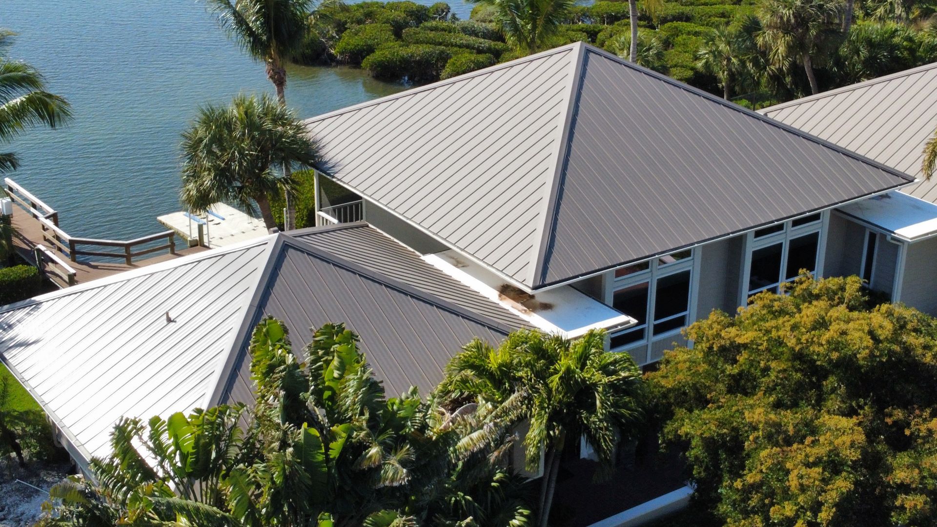Gray metal roof of a waterfront home with lush green foliage and blue water visible.