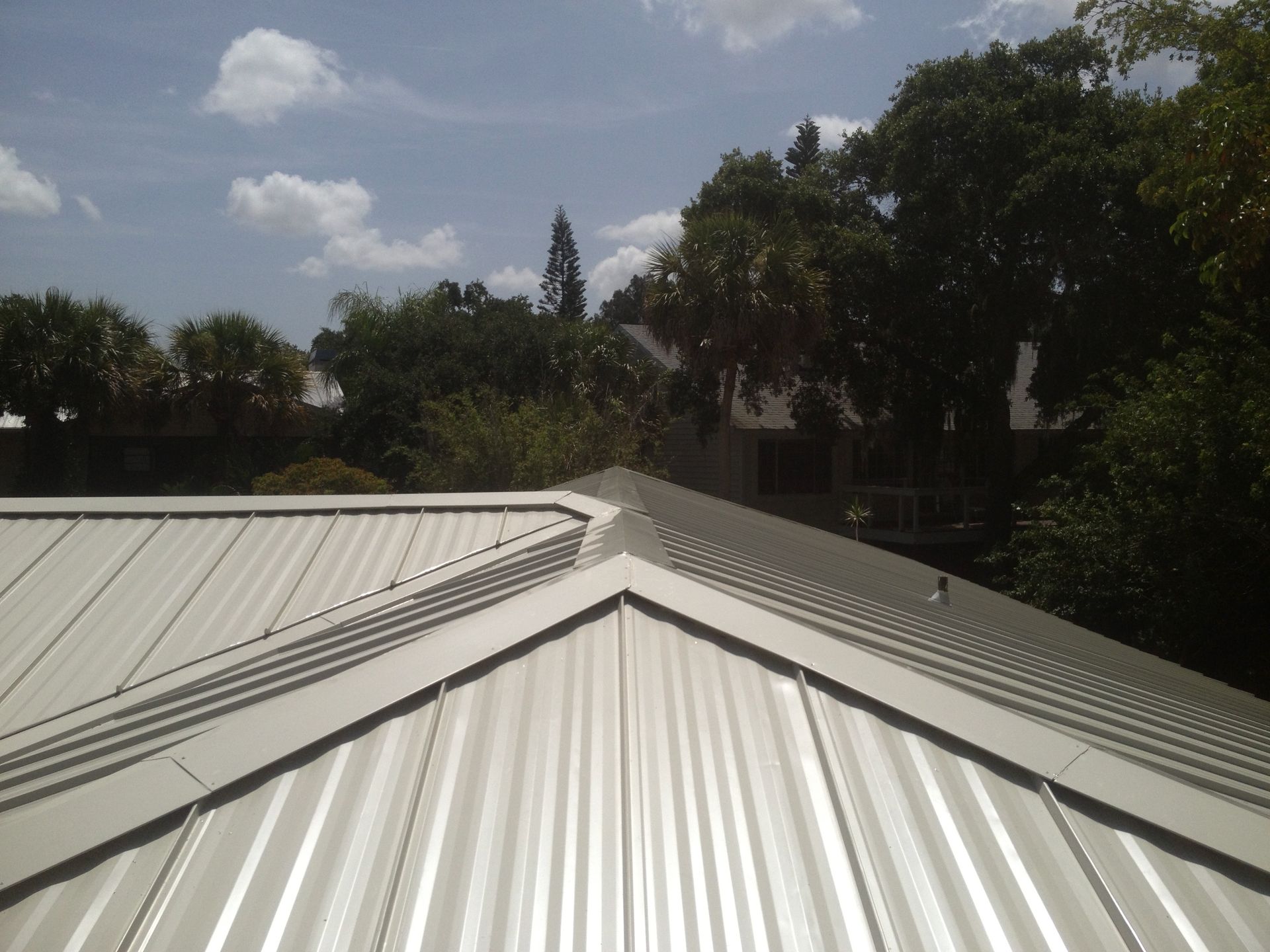 Metal roof with white panels, bright sunlight, trees and cloudy sky in background.