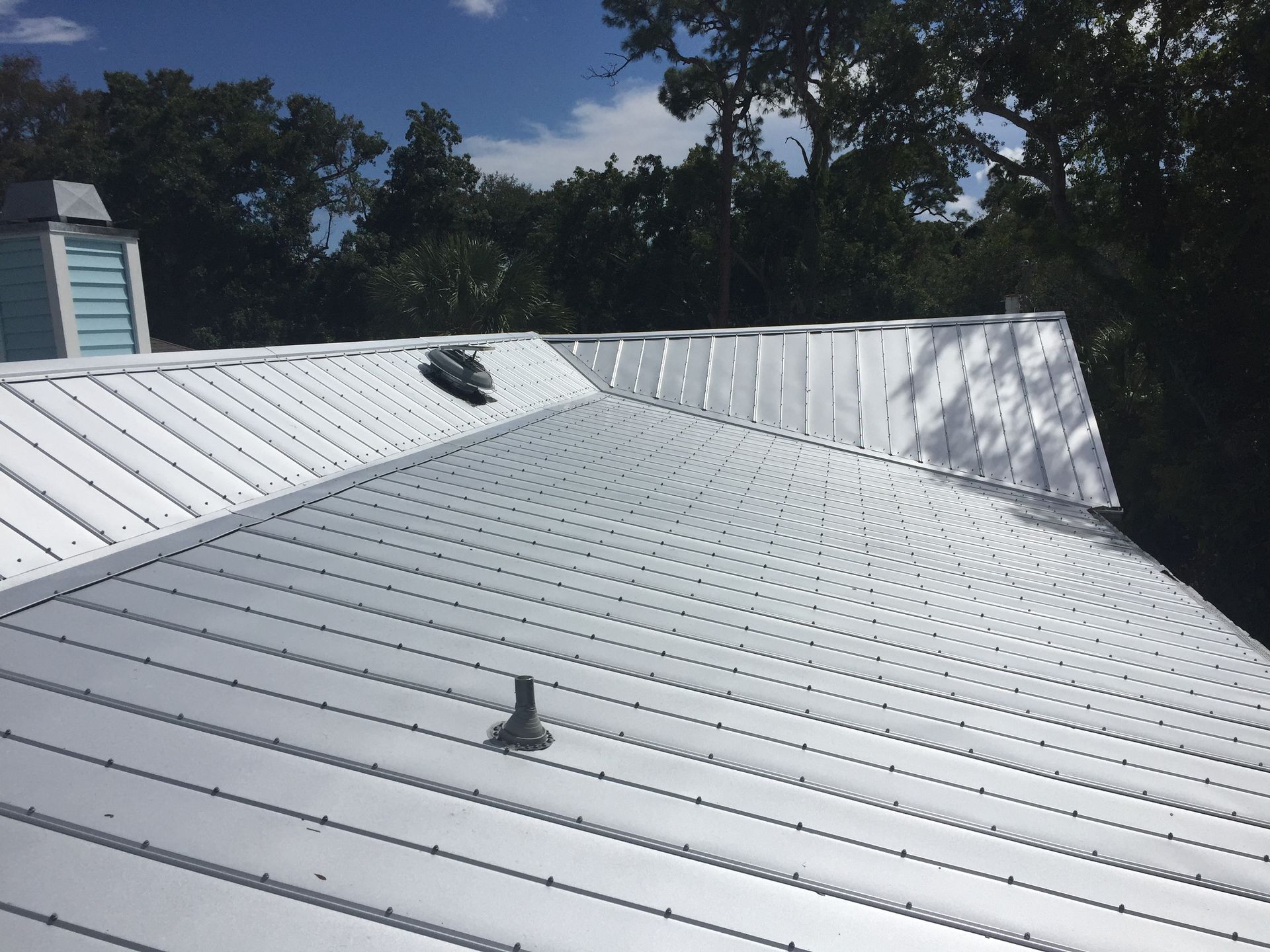 Metal roof with a vent and a pipe, surrounded by trees and a blue sky.