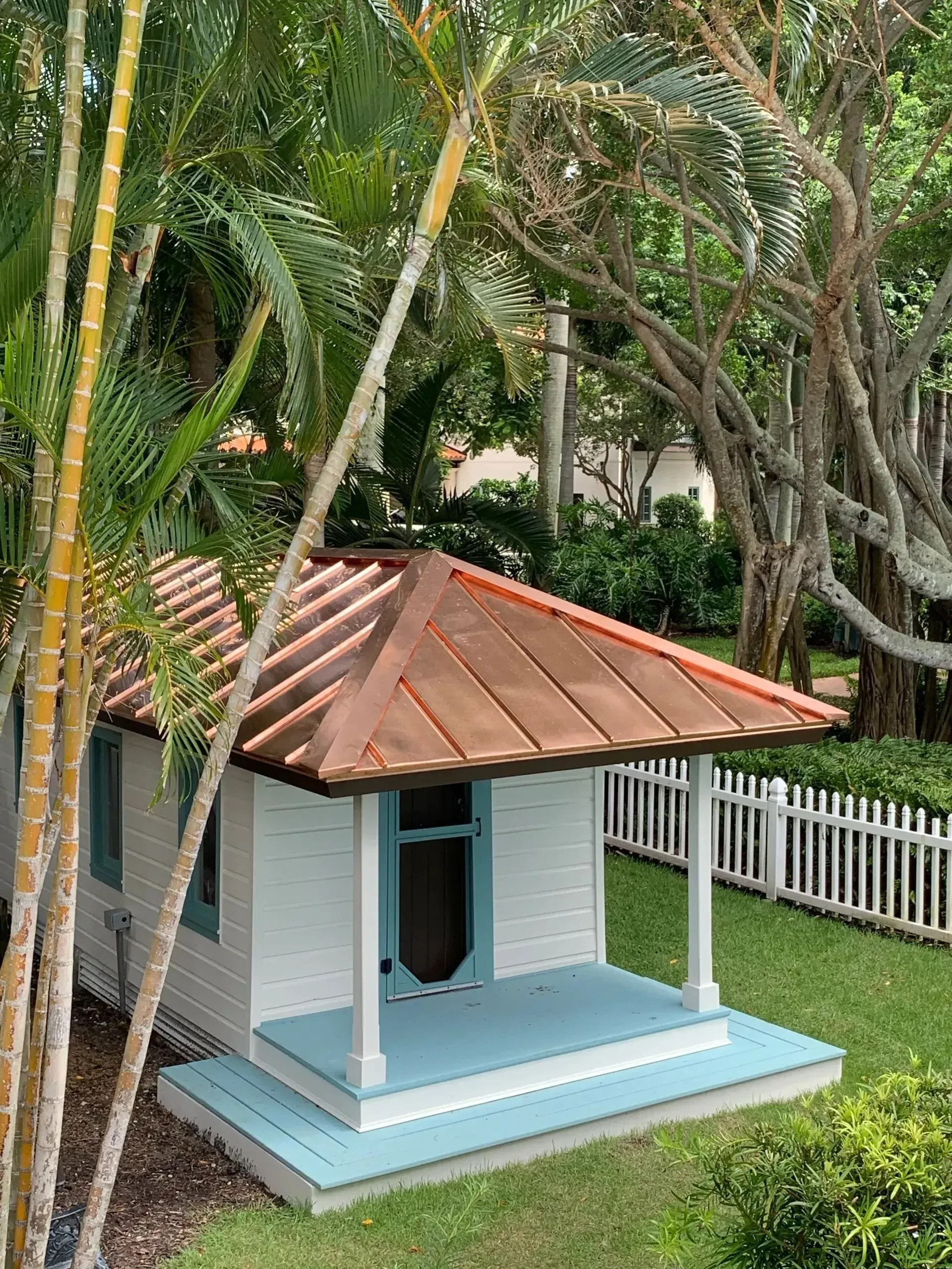 Doghouse with copper roof, blue accents, and white picket fence in a green yard.