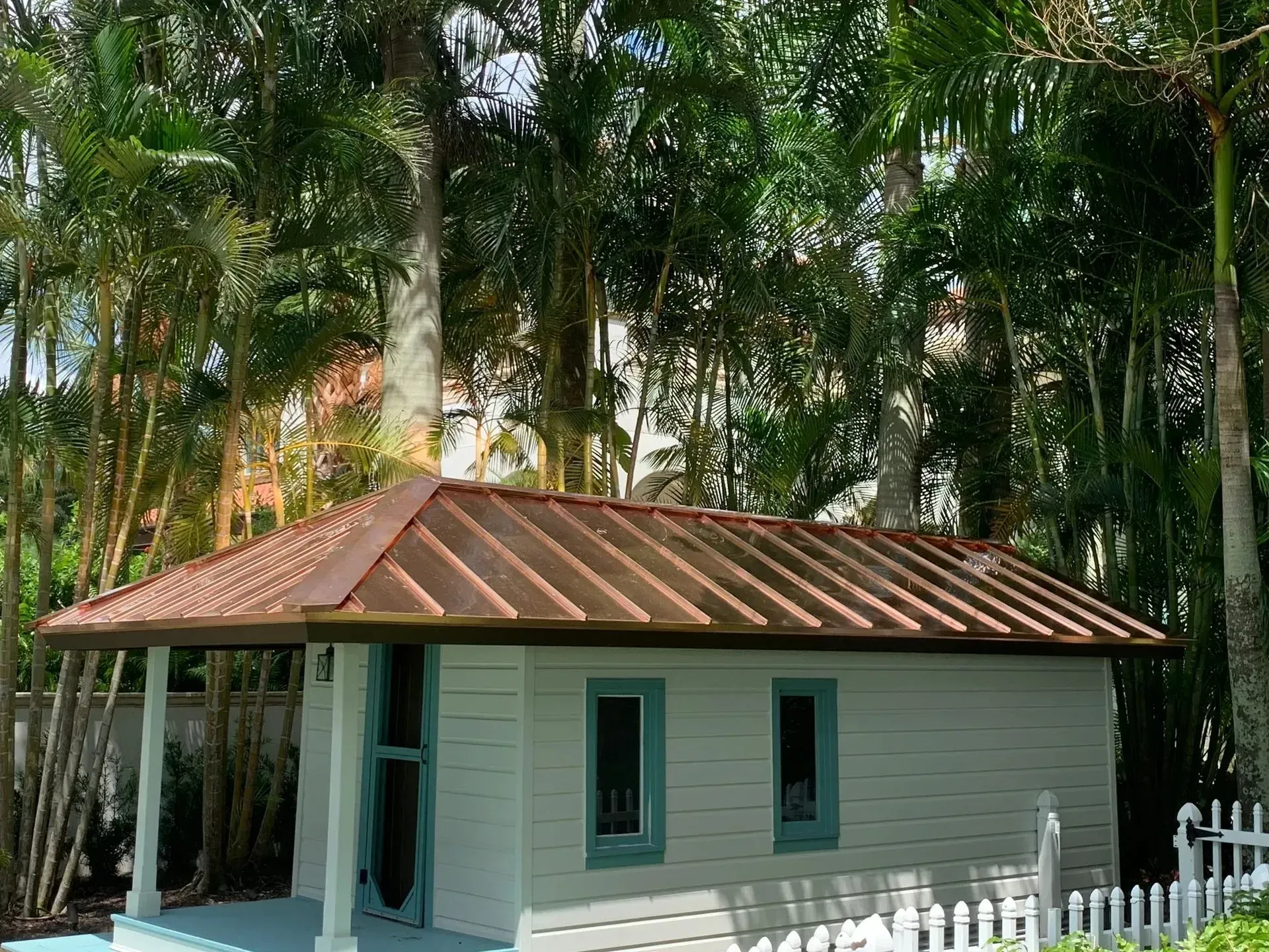 Small, light blue-grey building with a copper-colored roof and teal window frames, set among trees and a white picket fence.