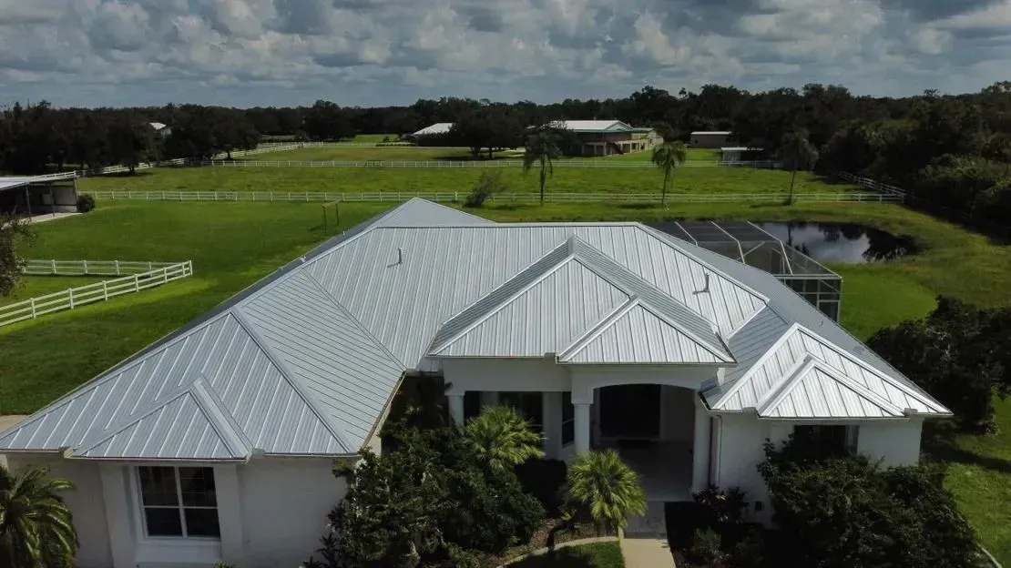 White house with a silver metal roof, surrounded by green grass, trees, and a pond.