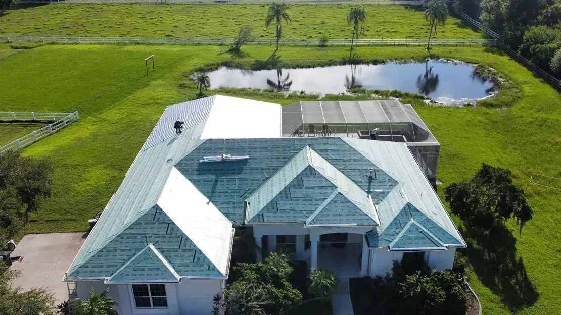 Aerial view of a white house with a teal-colored roof surrounded by green grass, trees, and a pond.