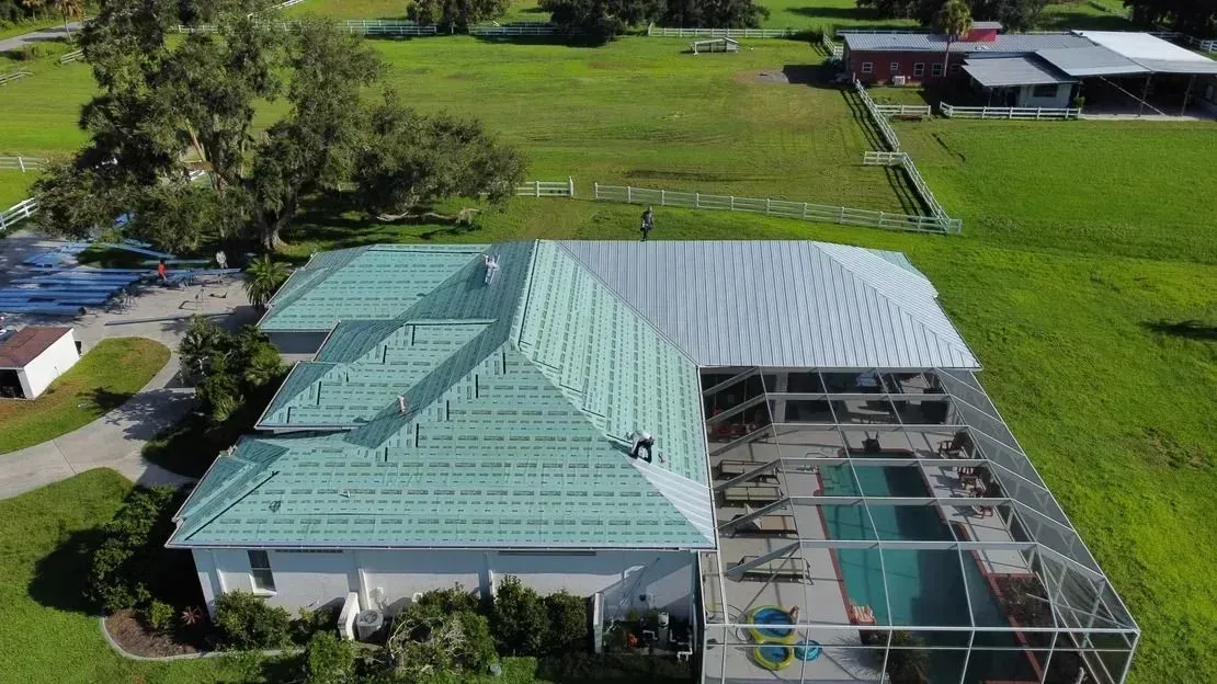 Aerial view of a house with a green roof, attached screened-in pool, and surrounding green lawn.