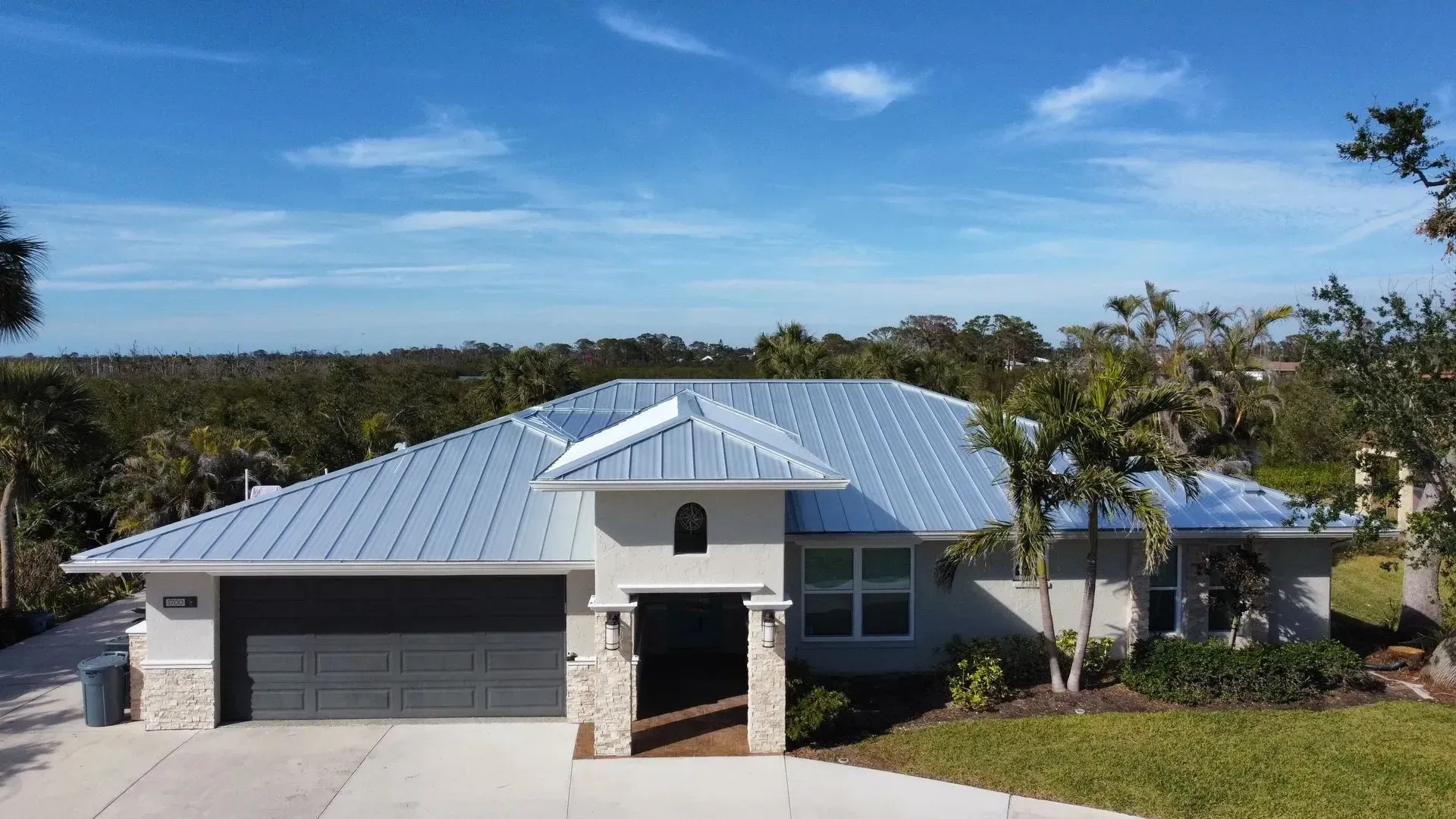 Modern home with a blue sky, metal roof, gray garage door, and tropical landscaping.