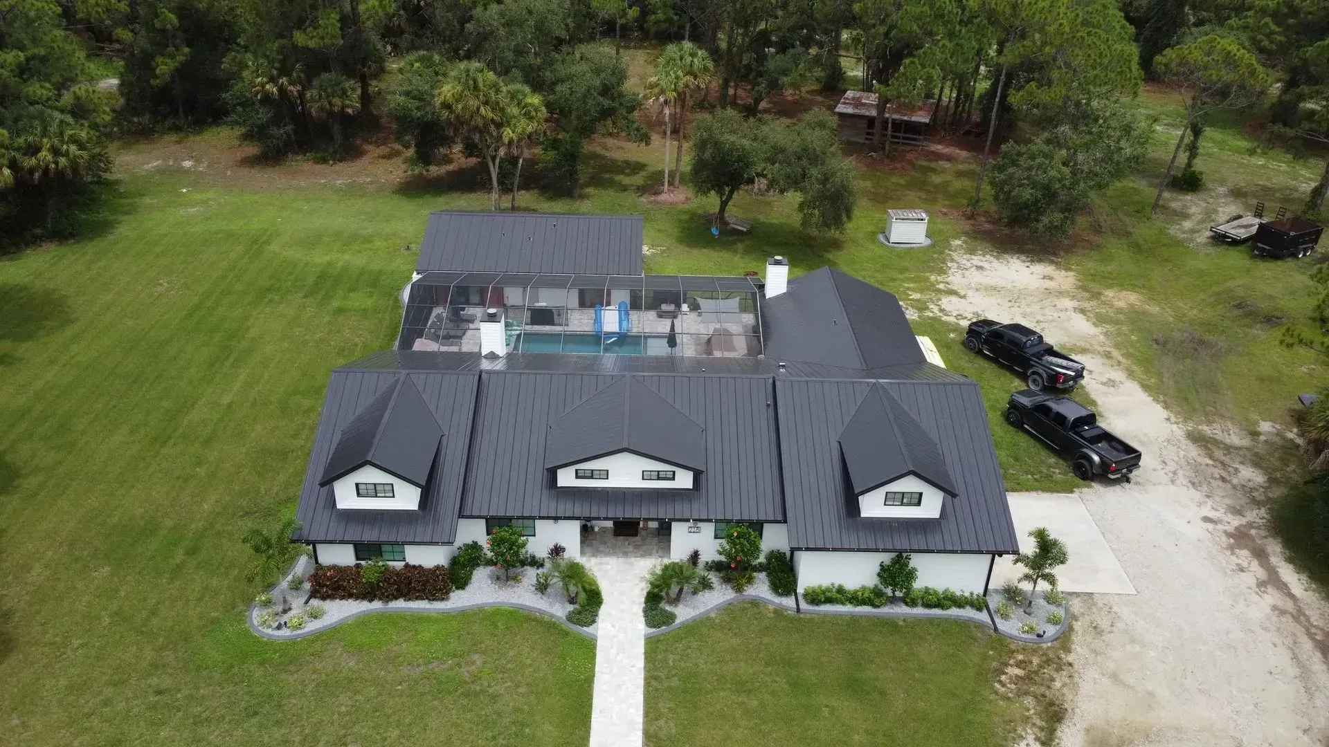 Aerial view of a white house with a dark roof and a pool. Black trucks parked outside on a gravel driveway.