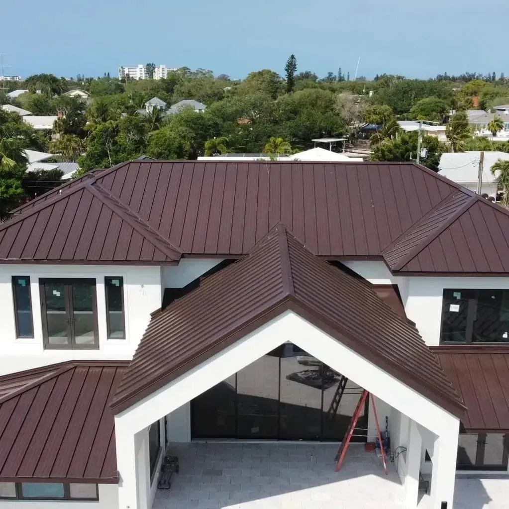 A brown metal roof on a white house, seen from above. Trees and other houses are in the background.