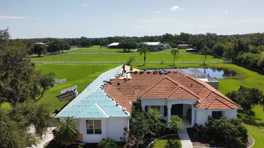 Roofers working on a house with a teal section and terracotta tiles, set in a green field with a pond.