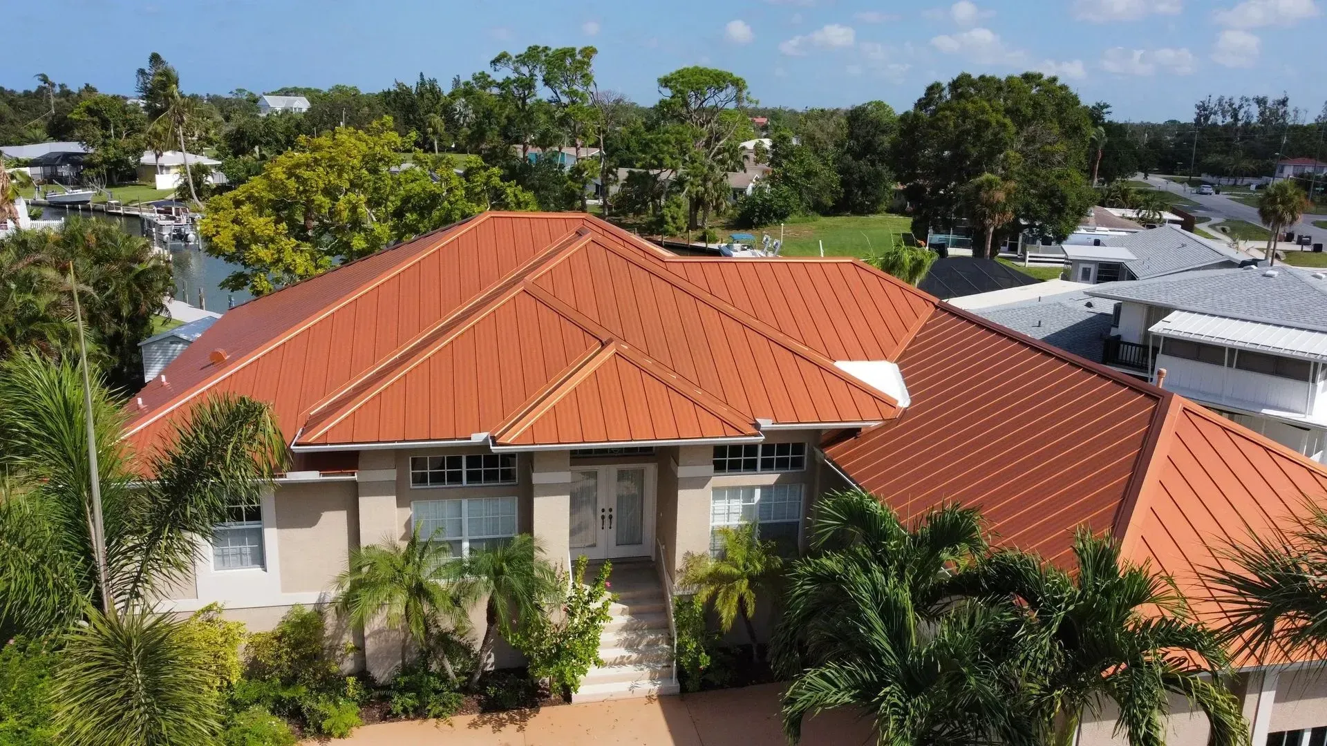 House with orange metal roof, surrounded by palm trees and other greenery in a sunny setting.