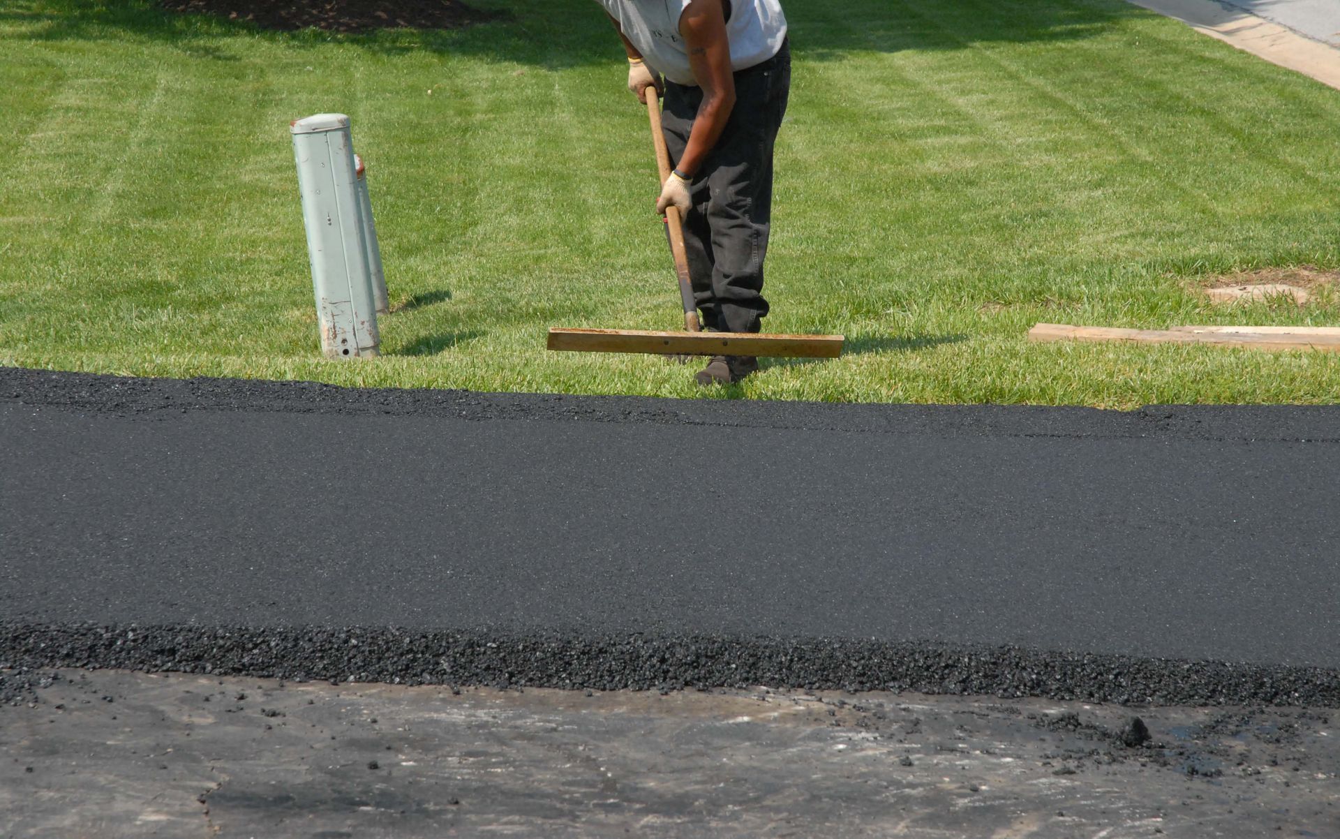 Worker paving a driveway, smoothing fresh black asphalt beside a green lawn.