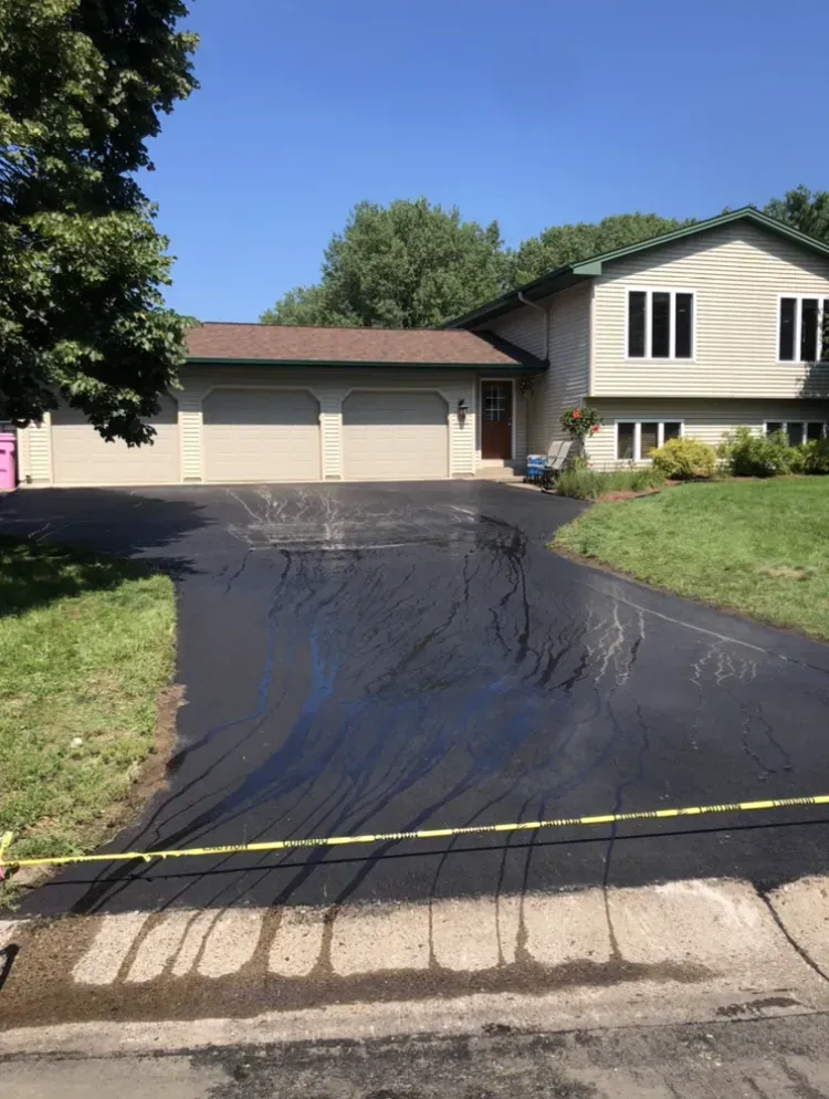 Fresh black asphalt driveway leading to a suburban house with two-car garage under a clear blue sky