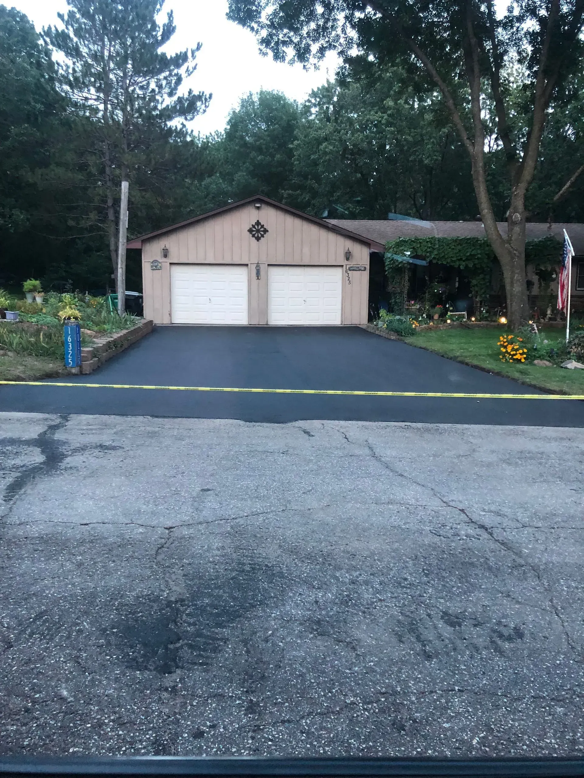 Driveway leading to a beige two-car garage surrounded by trees and flowers at dusk