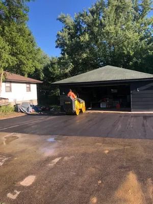 Worker using a yellow road machine on a freshly paved driveway beside a garage and house