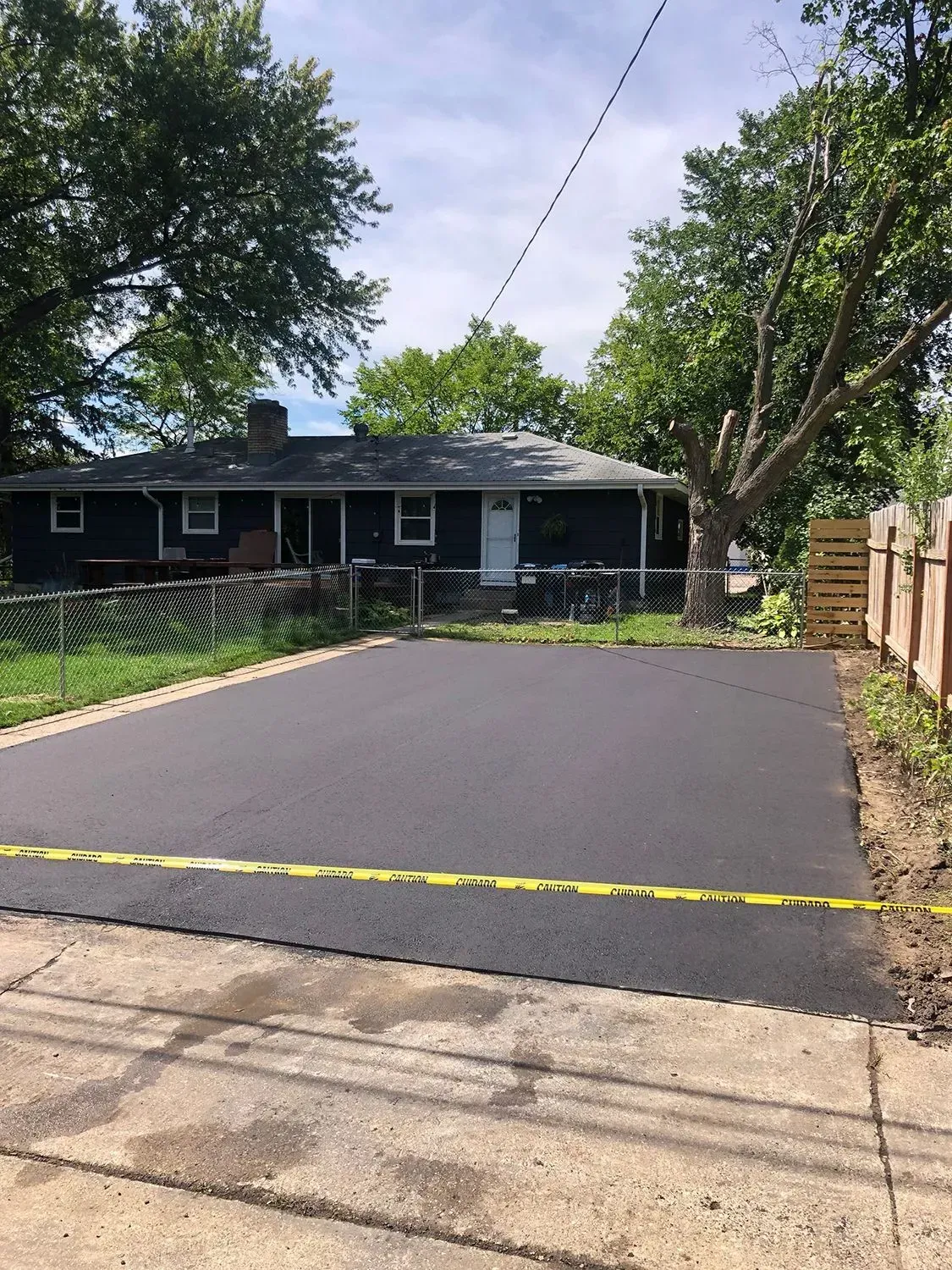 Fresh black asphalt driveway behind yellow caution tape, in front of a house and trees